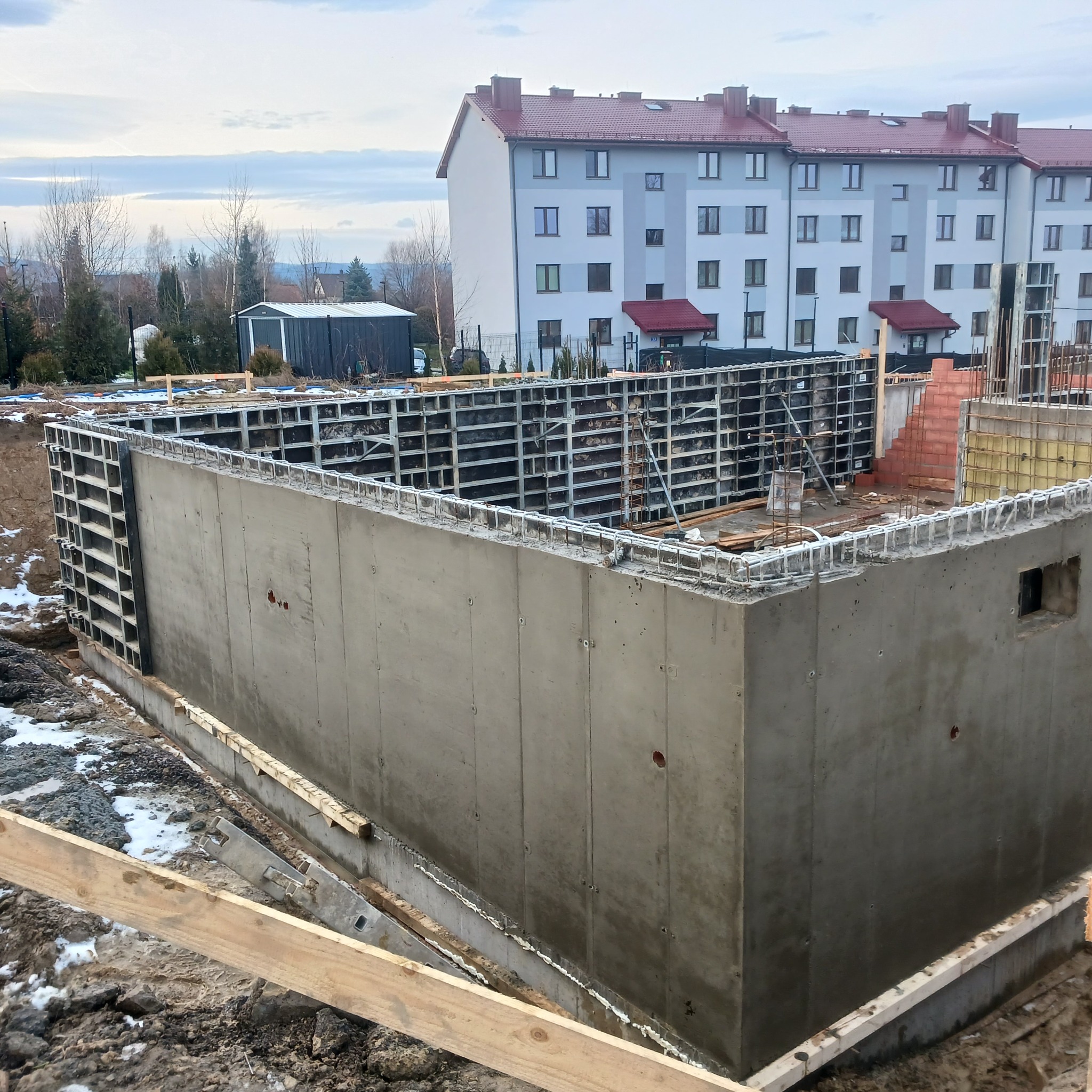 Betonowy fundament z szalunkiem, widoczne zbrojenie i otwór wentylacyjny, w tle budynek mieszkalny