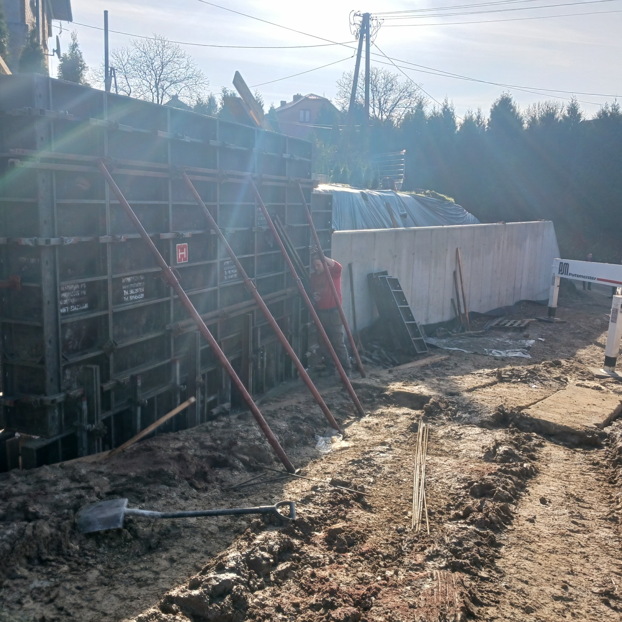 Szare szalunki podtrzymywane czerwonymi stemplami, pracownik w czerwonej koszuli wewnątrz konstrukcji, widoczny betonowy mur z logo Putzmeister, łopata w błocie, słoneczny dzień.