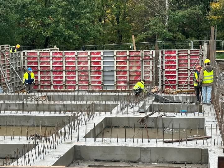 Wykopany i zalany betonem fundament z wystającymi prętami zbrojeniowymi, widoczne szalunki w kolorze czerwonym, pracownicy w odblaskowych kamizelkach i kaskach na budowie.