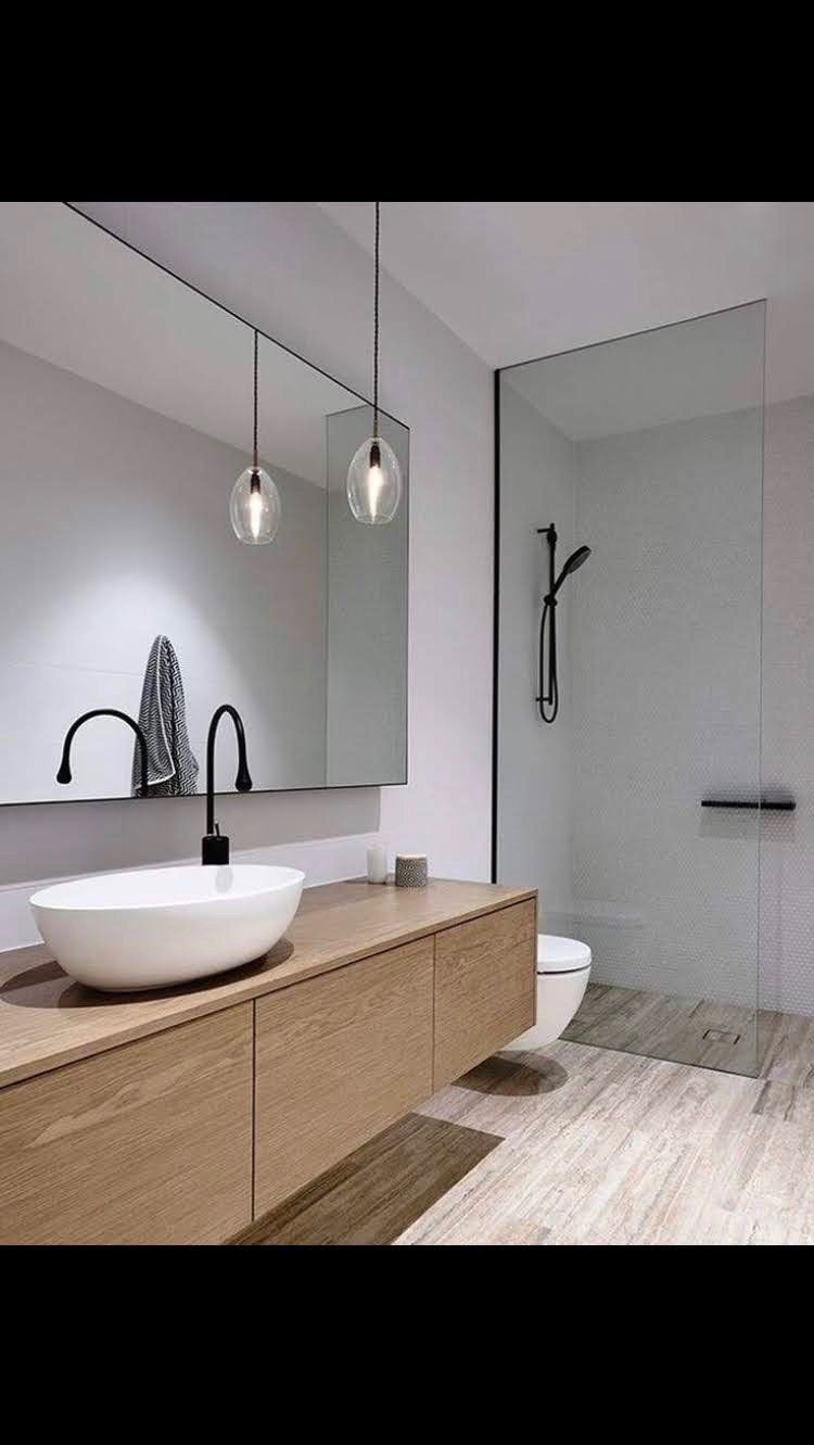 Nowoczesna łazienka z drewnianą szafką pod umywalką, owalną białą umywalką nablatową, czarną armaturą, dużym lustrem bez ramy, wiszącymi lampami w szklanych kloszach i kabiną prysznicową ze szklaną...