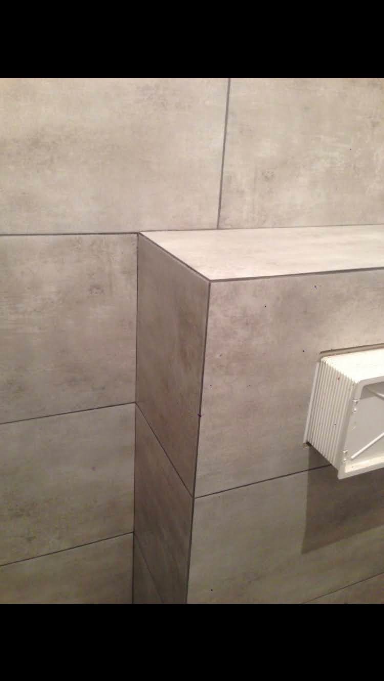 Wykończony fragment łazienki z szarymi płytkami imitującymi beton, widoczna narożna półka obłożona tymi samymi płytkami i biała kratka wentylacyjna.