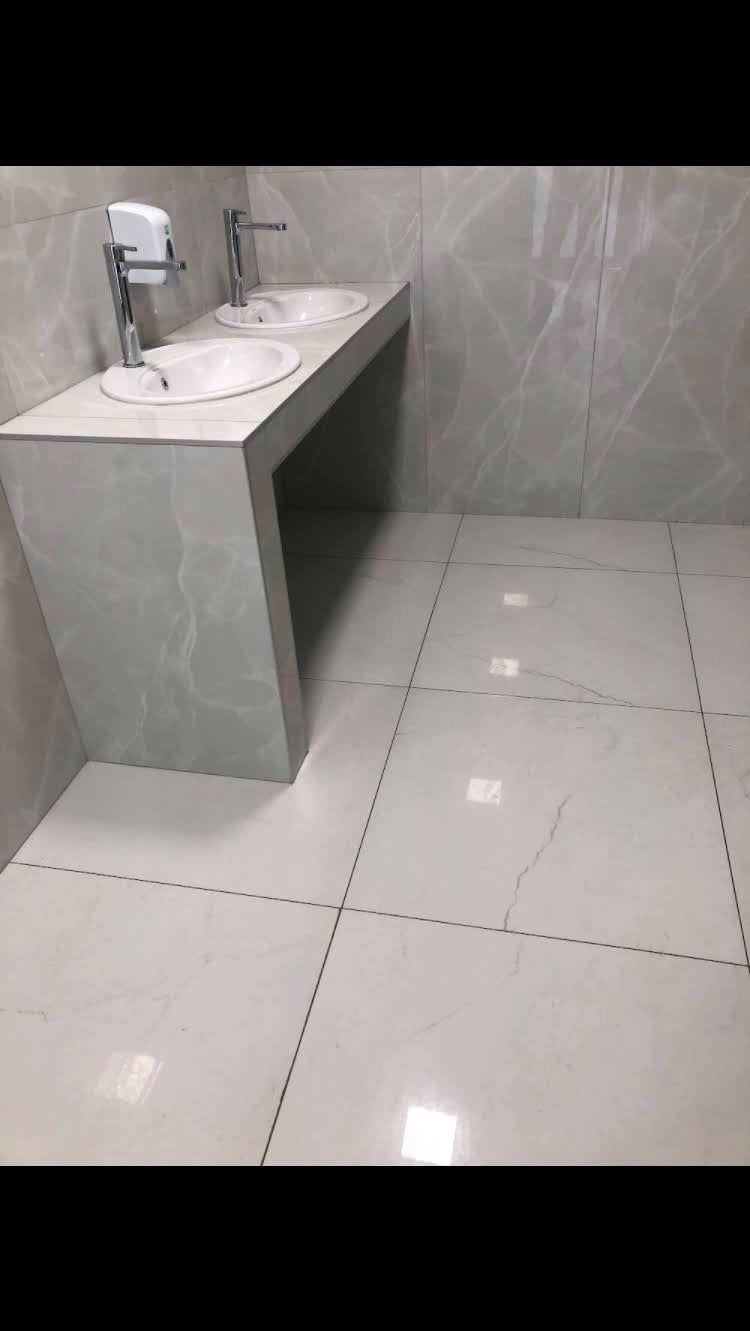 Dwie białe umywalki z chromowanymi kranami na blacie z płytek imitujących marmur, dopasowane do płytek na ścianach i podłodze, z dozownikiem mydła w tle.