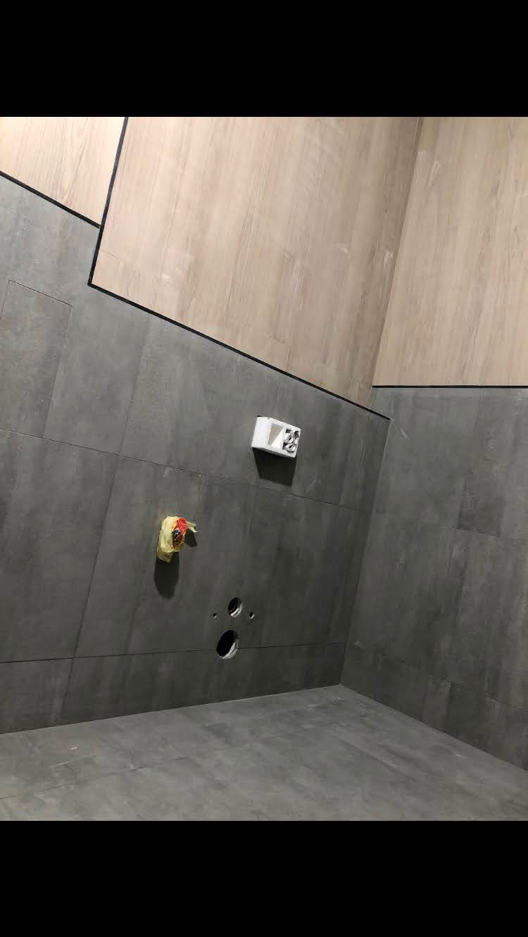 Wnętrze łazienki w trakcie wykańczania: szare płytki na ścianach i podłodze, drewnopodobne panele na górnej części ścian, widoczne otwory na instalacje, zabezpieczone rury.