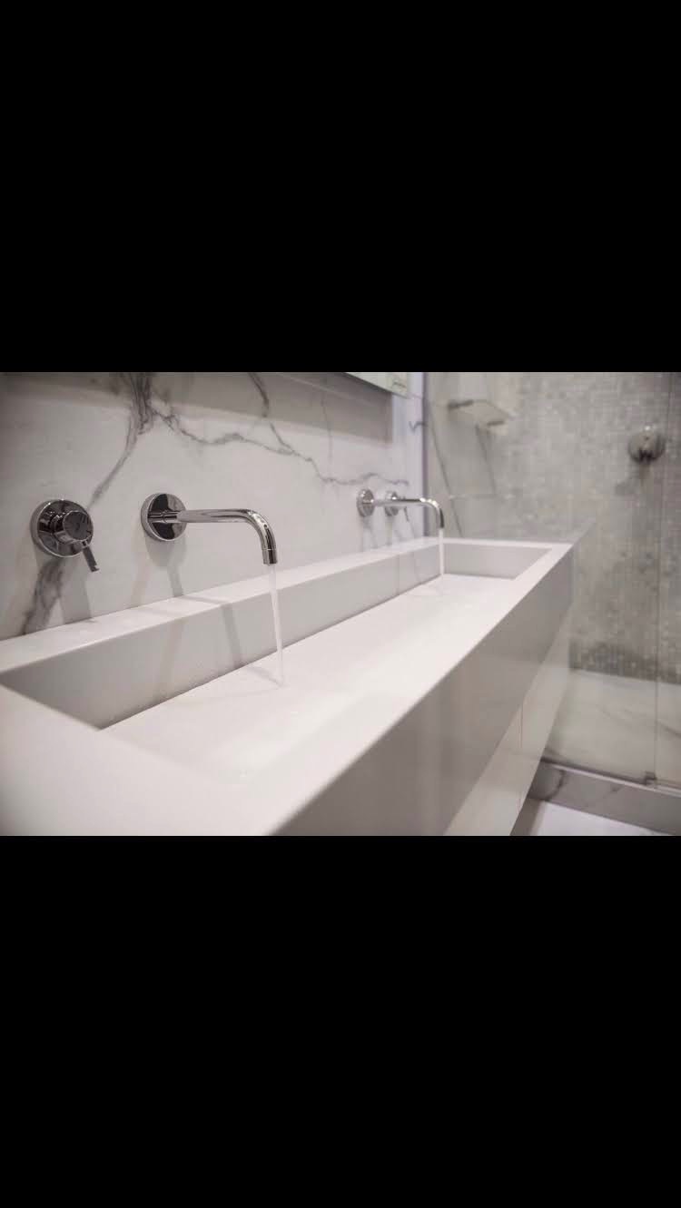 Nowoczesna, podwójna umywalka z białego kamienia na tle marmurowej ściany i szklanej kabiny prysznicowej, chromowane baterie z wypływającą wodą.