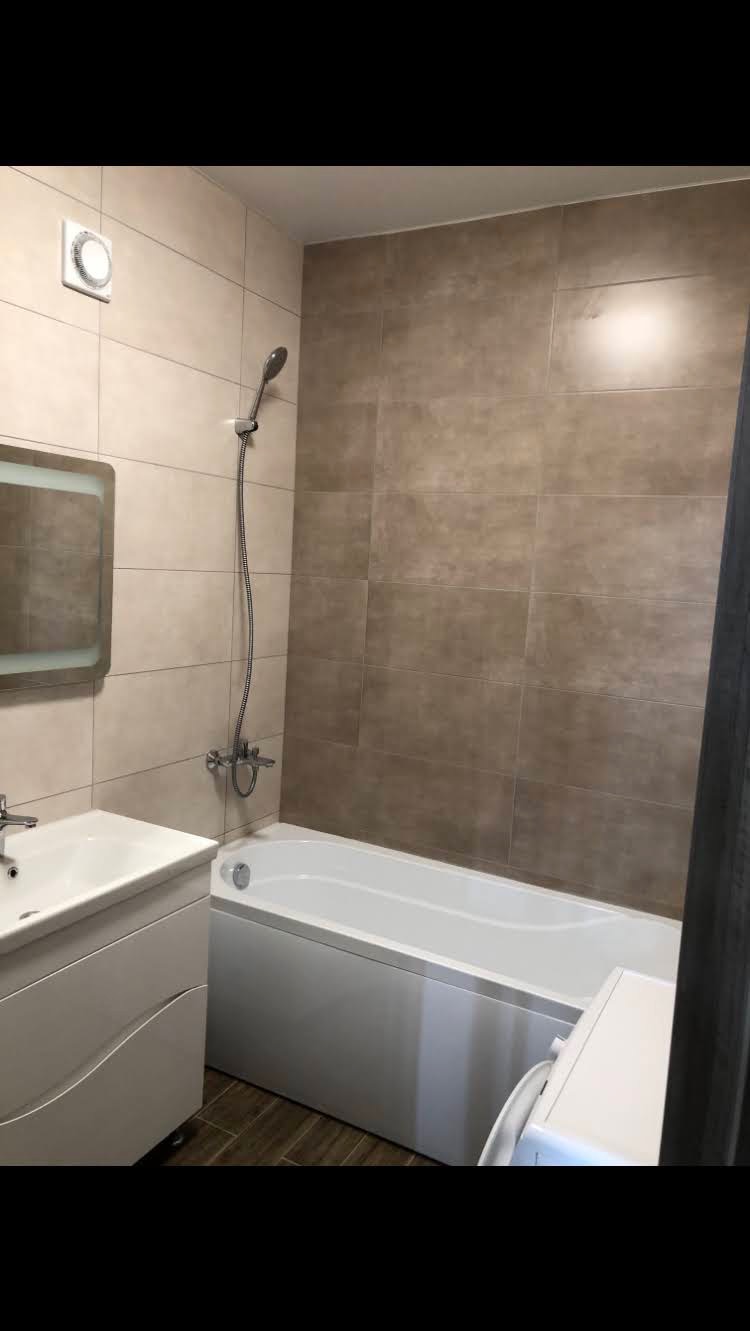 Wykończona łazienka z beżowymi płytkami na ścianach, białą wanną i umywalką z szafką, widoczny fragment pralki z boku.