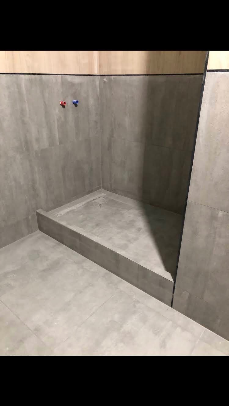 Przygotowany brodzik z odpływem liniowym w łazience wyłożonej szarą, matową glazurą, widoczne wystające rury ciepłej i zimnej wody.