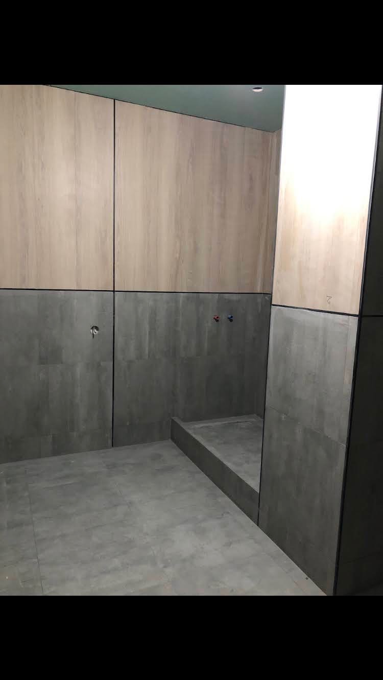 Wnętrze łazienki z wykończonymi ścianami częściowo panelami imitującymi drewno, a częściowo szarymi płytkami, widoczny fragment brodzika i wystające przyłącza wodne.