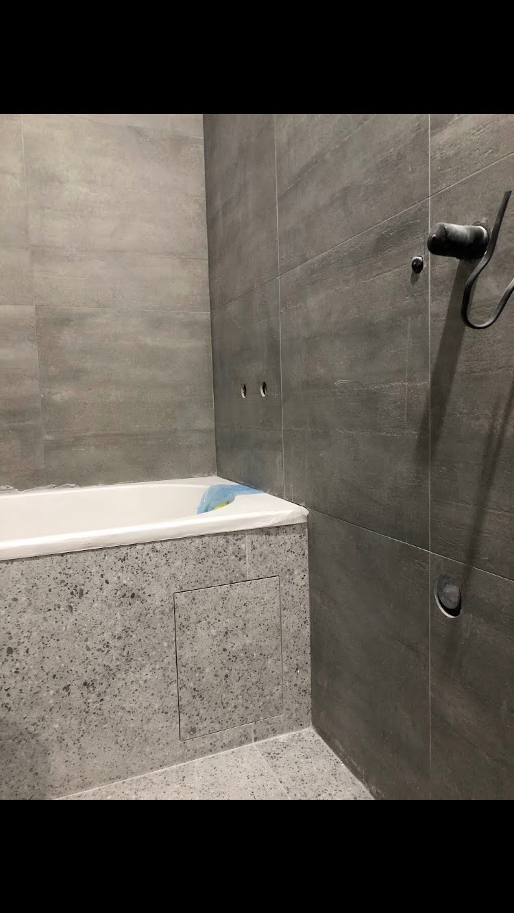 Nowoczesna łazienka z szarymi płytkami imitującymi beton, białą wanną zabezpieczoną folią i ukrytą klapą rewizyjną w obudowie wanny.