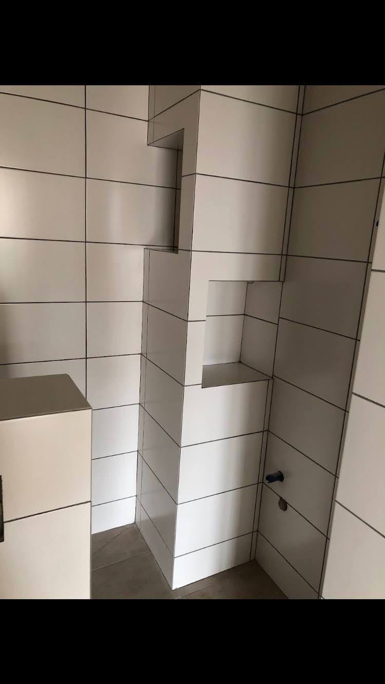 Białe, prostokątne płytki ceramiczne na ścianie z wnęką w kształcie litery L, widoczny fragment szafki i szara podłoga