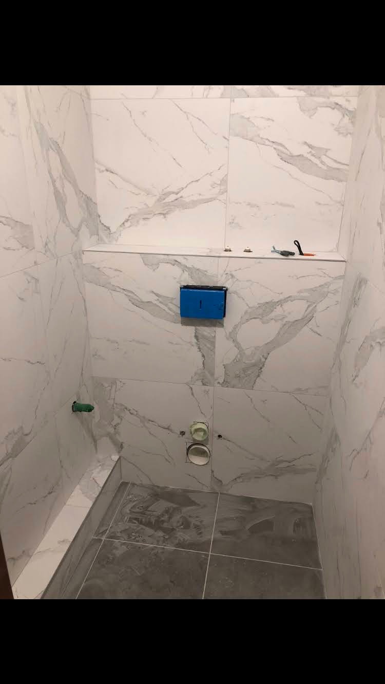 Łazienka w trakcie wykańczania: marmurowe płytki na ścianach, szare płytki na podłodze, widoczne instalacje hydrauliczne i narzędzia glazurnicze na półce.