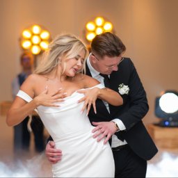 tojasun fotografia Sandra Stefańska - Para młoda w tańcu podczas wesela, ona w białej sukni z odkrytymi ramionami, on w czarnym garniturze z butonierką. Scena oświetlona reflektorami.