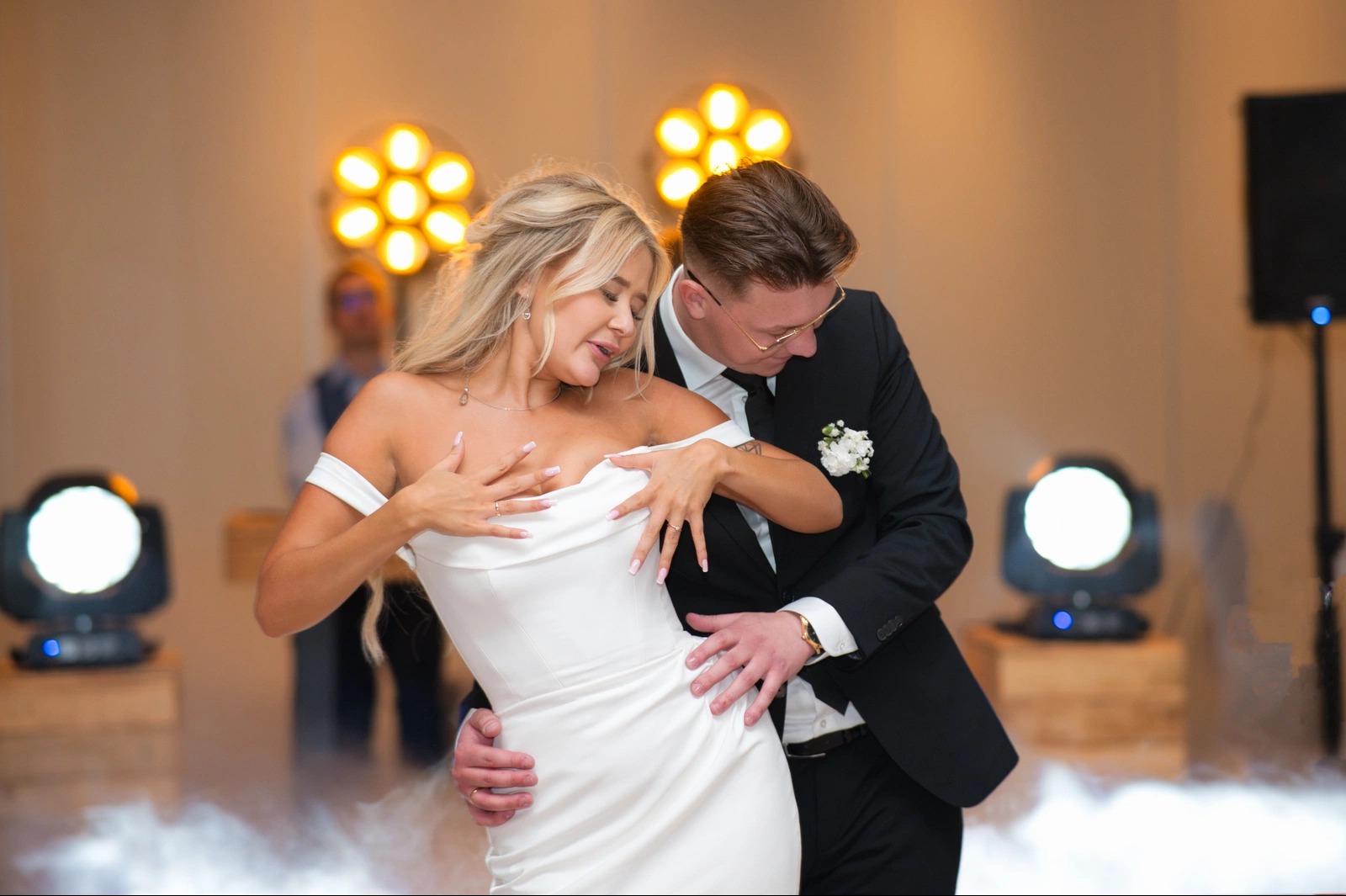 Para młoda w tańcu podczas wesela, ona w białej sukni z odkrytymi ramionami, on w czarnym garniturze z butonierką. Scena oświetlona reflektorami.