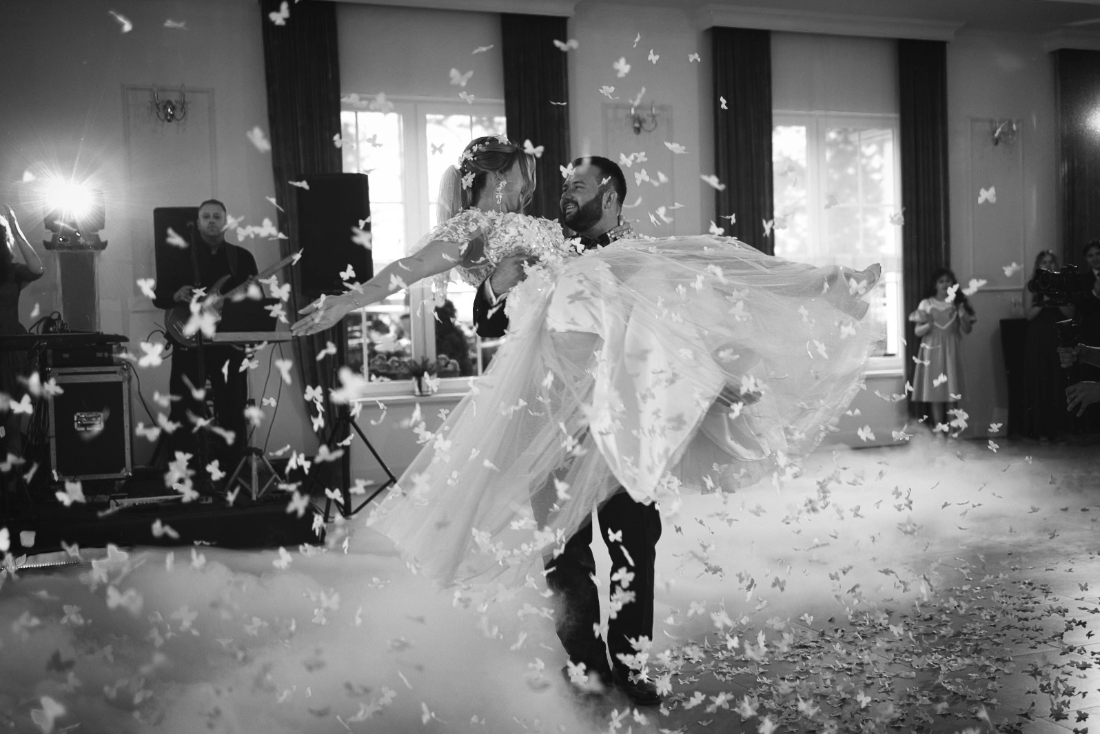 Czarno-białe ujęcie pary młodej podczas pierwszego tańca, unoszonej przez pana młodego, w otoczeniu konfetti w kształcie motyli i dymu na parkiecie.