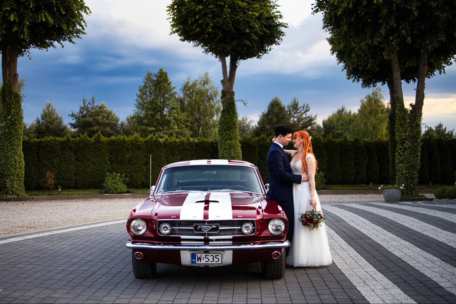 Para młoda obejmuje się przy czerwonym Fordzie Mustang z białym pasem na tle zielonego żywopłotu i drzew. Romantyczny nastrój, klasyczna elegancja.