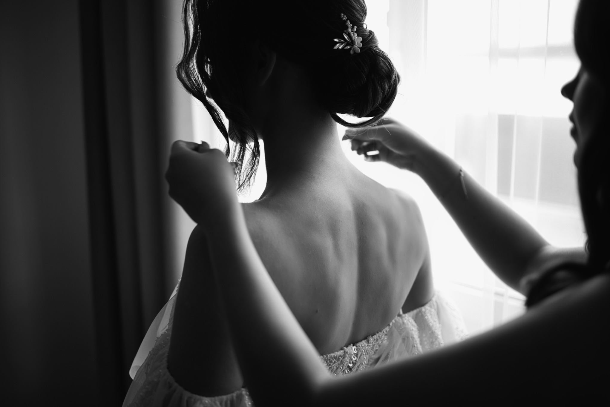 Czarno-białe ujęcie panny młodej z tyłu, gdy druhna zapina jej suknię ślubną przy oknie, delikatne upięcie włosów z ozdobą.