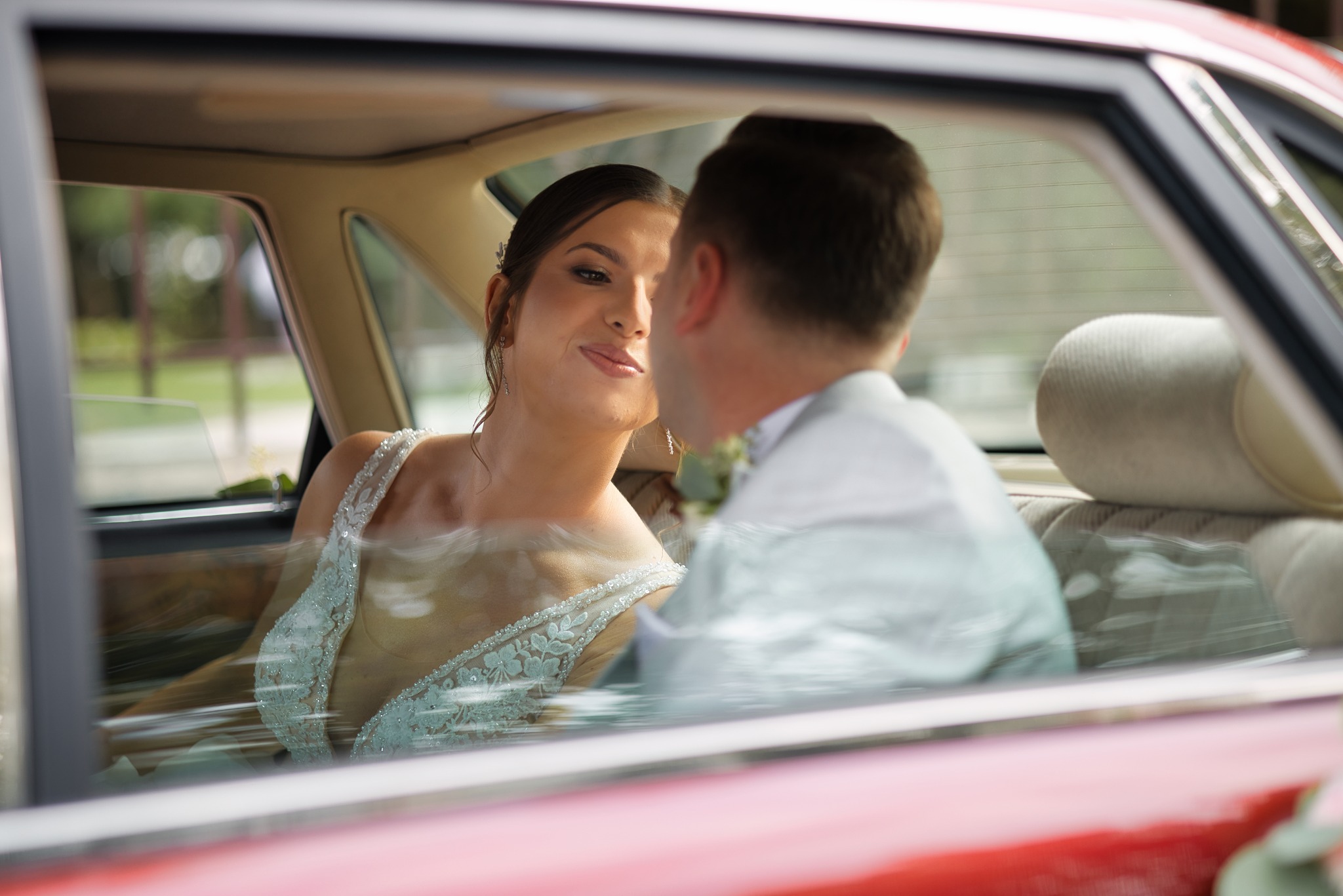 Panna młoda w sukni z koronką spogląda na pana młodego w samochodzie retro. Delikatne oświetlenie podkreśla ich emocje w dniu ślubu.