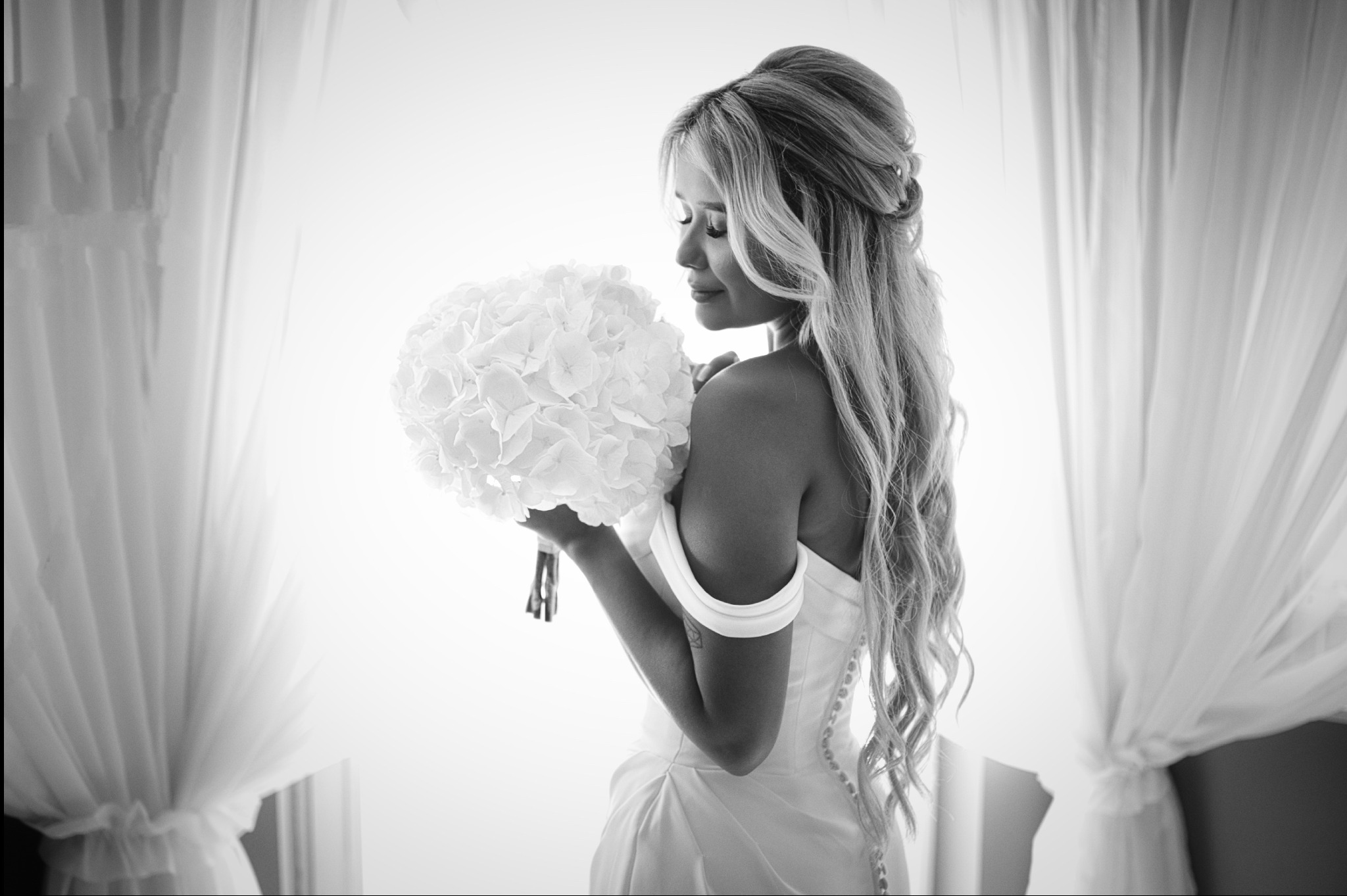 Elegancka panna młoda w sukni, z bukietem, w czarno-białej tonacji, stoi na tle okna z białymi zasłonami. Delikatne światło podkreśla fryzurę i detale sukni.