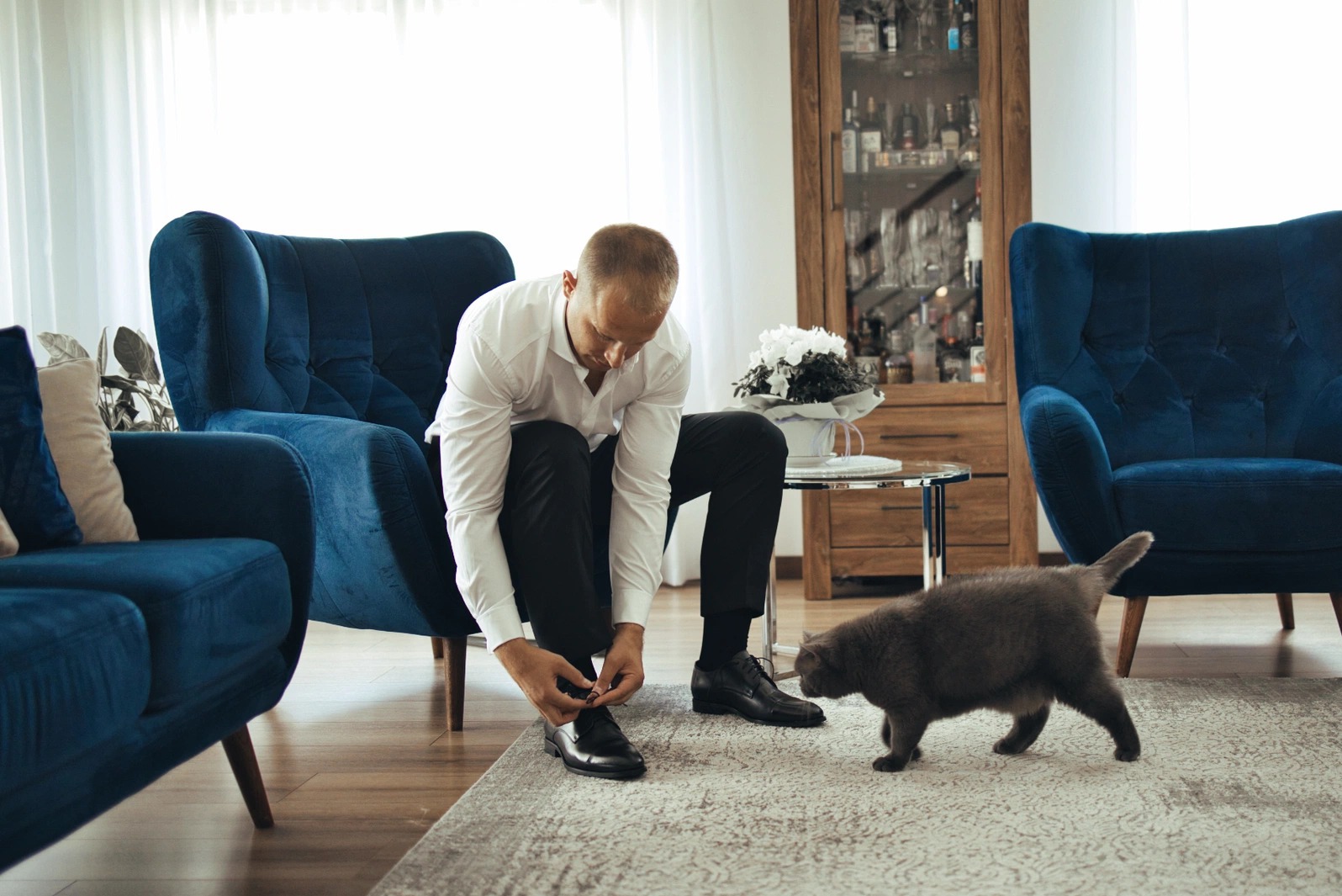Elegancki mężczyzna wiąże sznurówki butów, a obok przechadza się kot. Zdjęcie wykonano w Warszawie, w jasnym wnętrzu z niebieskimi fotelami.