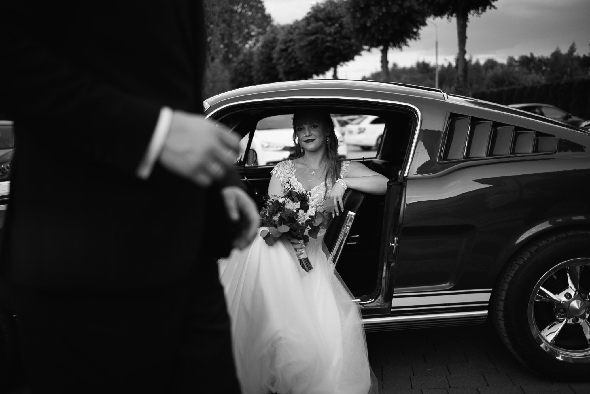 Czarno-białe zdjęcie panny młodej w sukni ślubnej, siedzącej w klasycznym samochodzie Mustang, trzymającej bukiet. W tle drzewa i inny samochód.