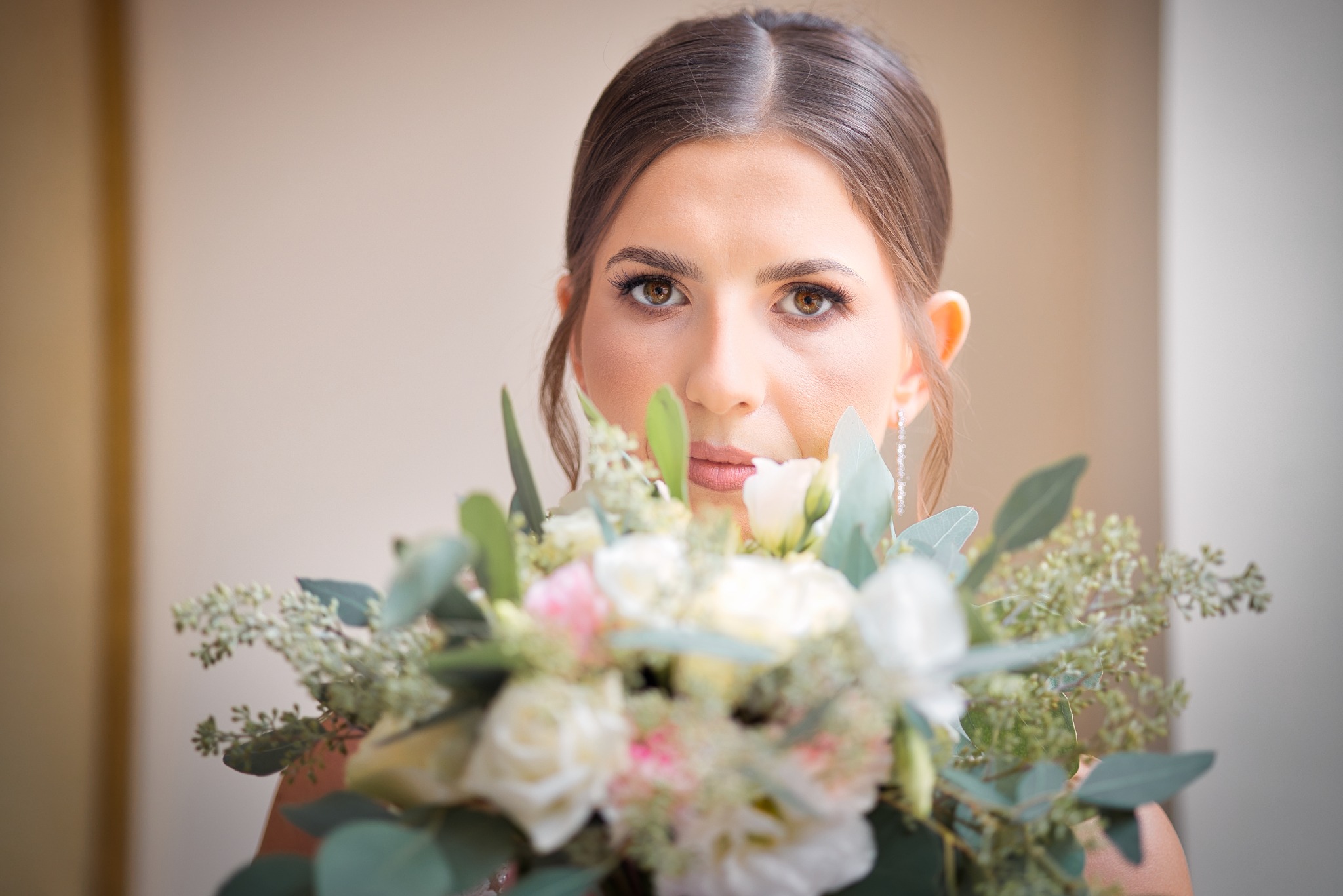 Portret panny młodej z bukietem ślubnym w dłoniach. Delikatny makijaż i elegancka fryzura, stonowane tło. Ujęcie centralne, skupione na twarzy i kwiatach.