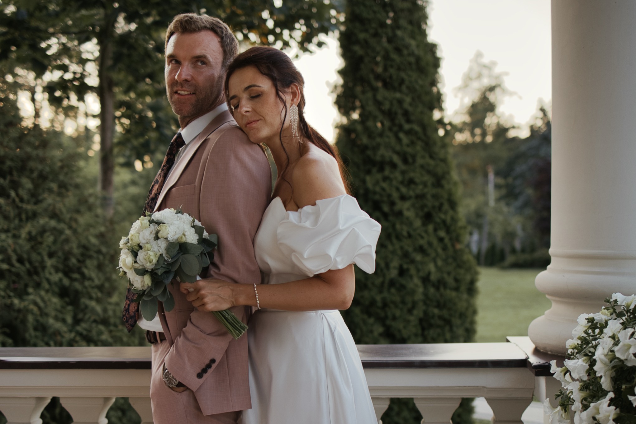 Elegancka para młoda w objęciach na balkonie. Pan w różowym garniturze, pani w białej sukni z bukietem. Romantyczna atmosfera, naturalne światło, zielone tło.