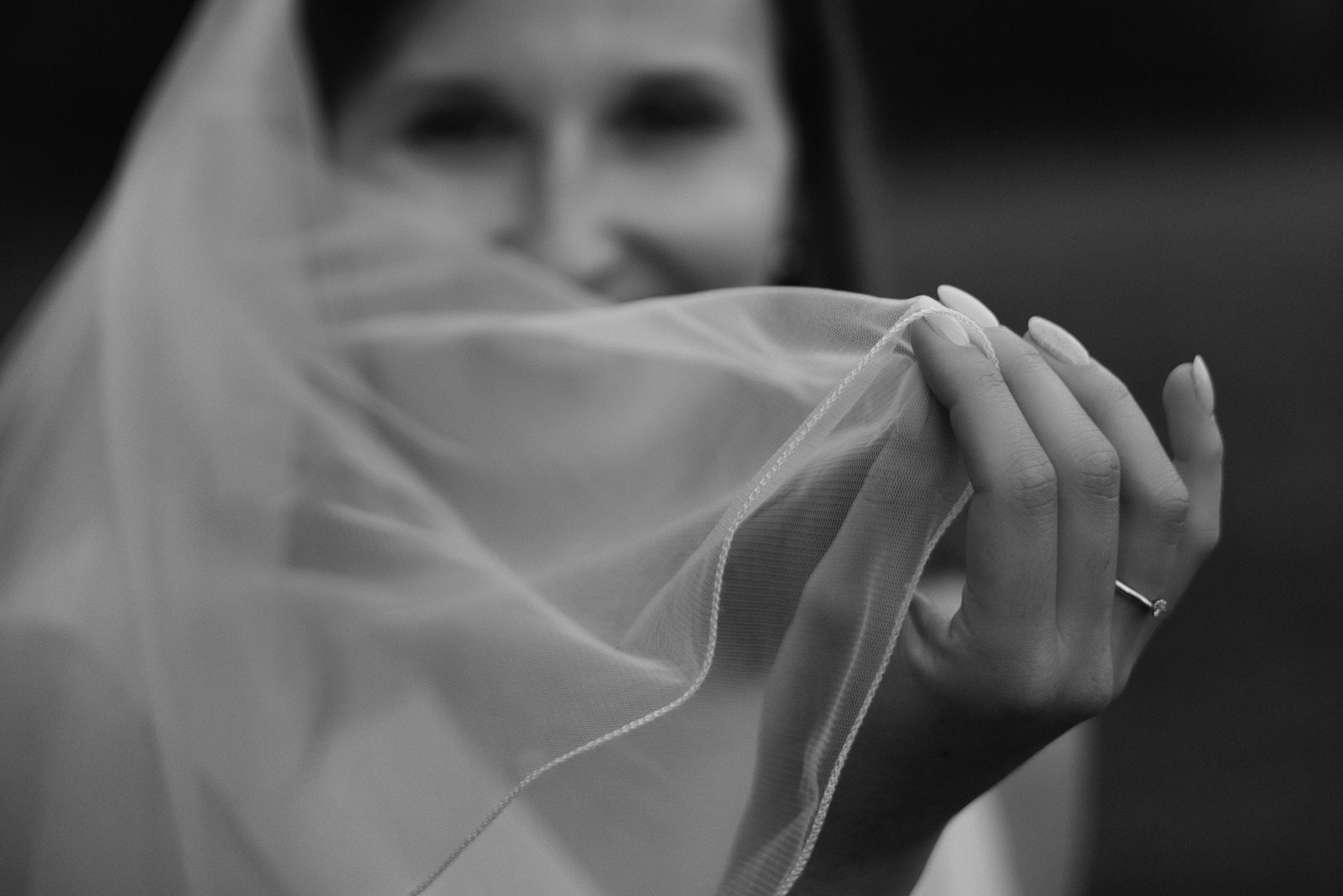 Czarno-białe zbliżenie dłoni panny młodej z pierścionkiem, trzymającej delikatny welon na tle zamazanej twarzy. Subtelne światło i detale.