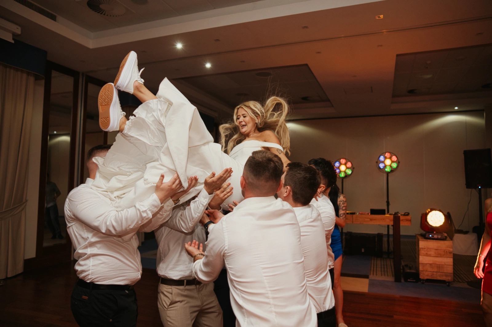 Radosna panna młoda w białej sukni i sportowych butach, uniesiona w górę przez grupę mężczyzn w białych koszulach podczas weselnego przyjęcia.