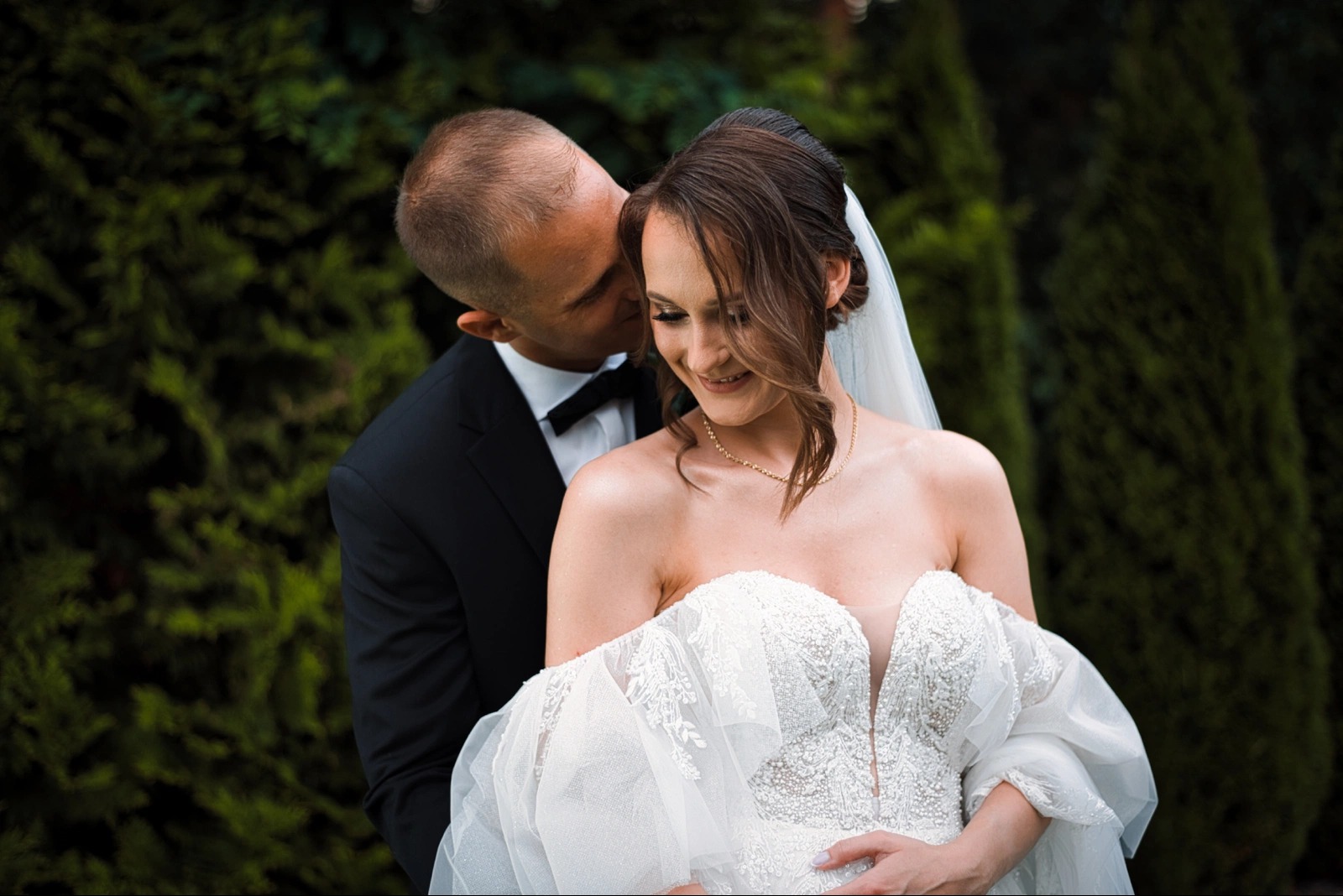 Uśmiechnięta panna młoda w koronkowej sukni, delikatnie obejmowana przez pana młodego na tle zielonych krzewów. Romantyczny moment ślubny.