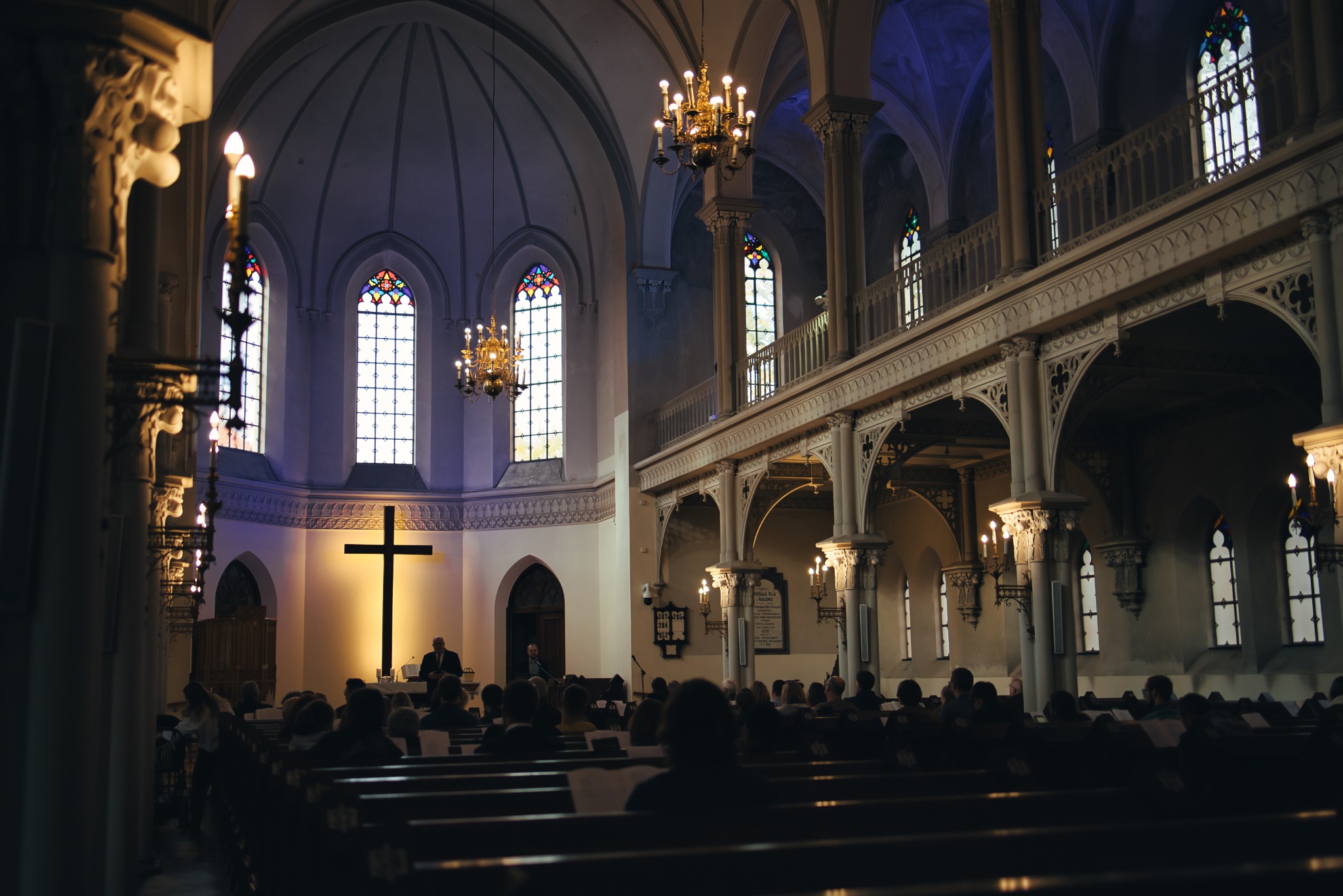 Wnętrze kościoła z wysokimi oknami witrażowymi, żyrandolami i rzędami ławek wypełnionych wiernymi podczas mszy. Na ścianie widoczny krzyż.