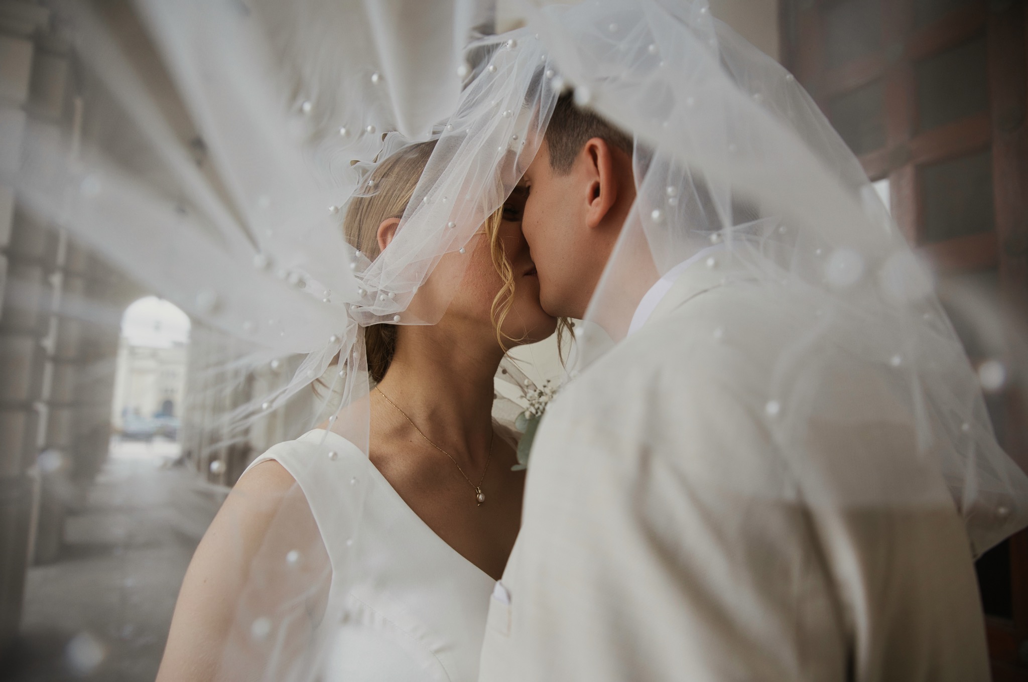 Pocałunek pary młodej pod welonem z perłami, w tle kolumnada. Romantyczne ujęcie z sesji ślubnej, delikatne światło podkreśla intymność chwili.