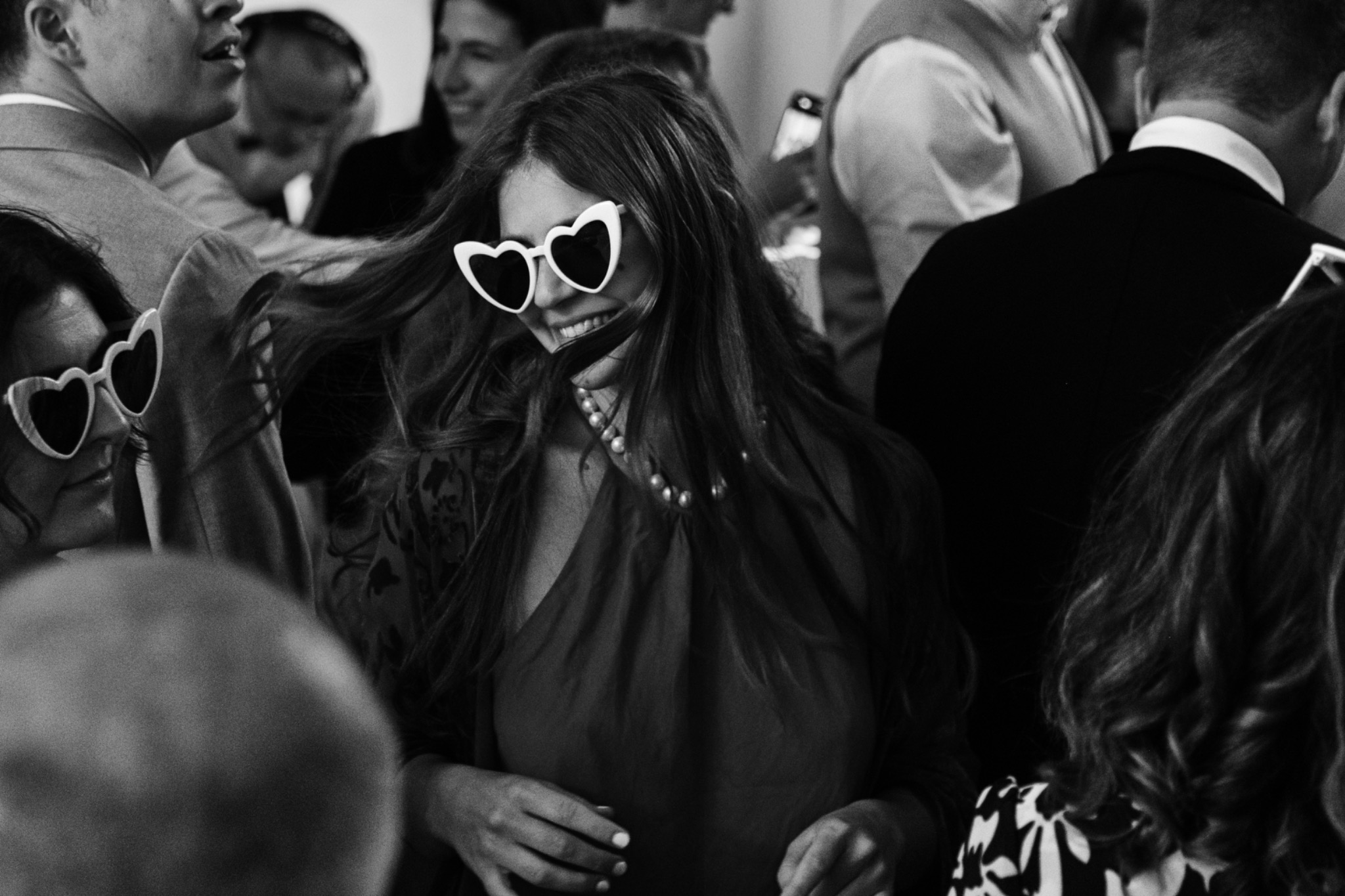 Czarno-białe zdjęcie uśmiechniętej kobiety w okularach w kształcie serc, otoczonej przez ludzi w tle, uchwycone w dynamicznym ruchu.