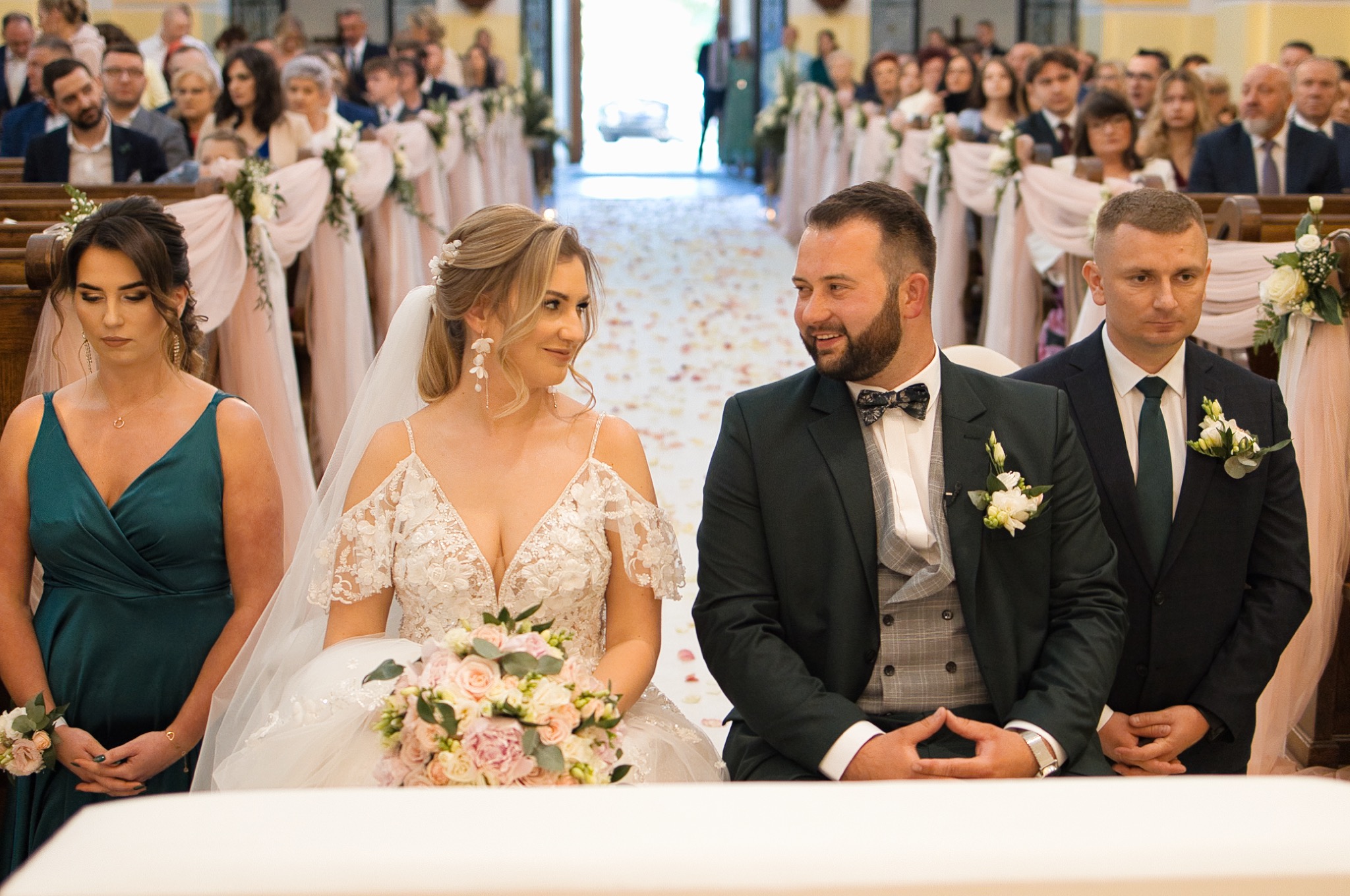 Para młoda podczas ceremonii ślubnej w kościele. Panna młoda w białej sukni z bukietem, pan młody w ciemnozielonym garniturze. W tle goście siedzący w ławkach.