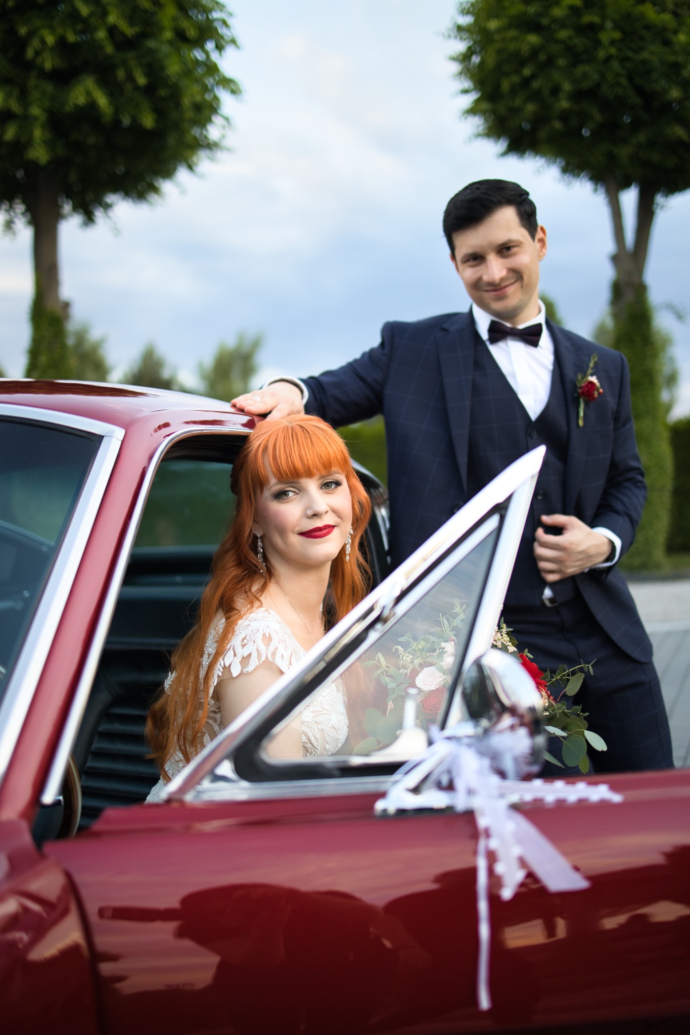 Para młoda pozuje przy czerwonym, klasycznym samochodzie. Panna młoda z rudymi włosami siedzi w aucie, a pan młody w granatowym garniturze opiera się o dach pojazdu.