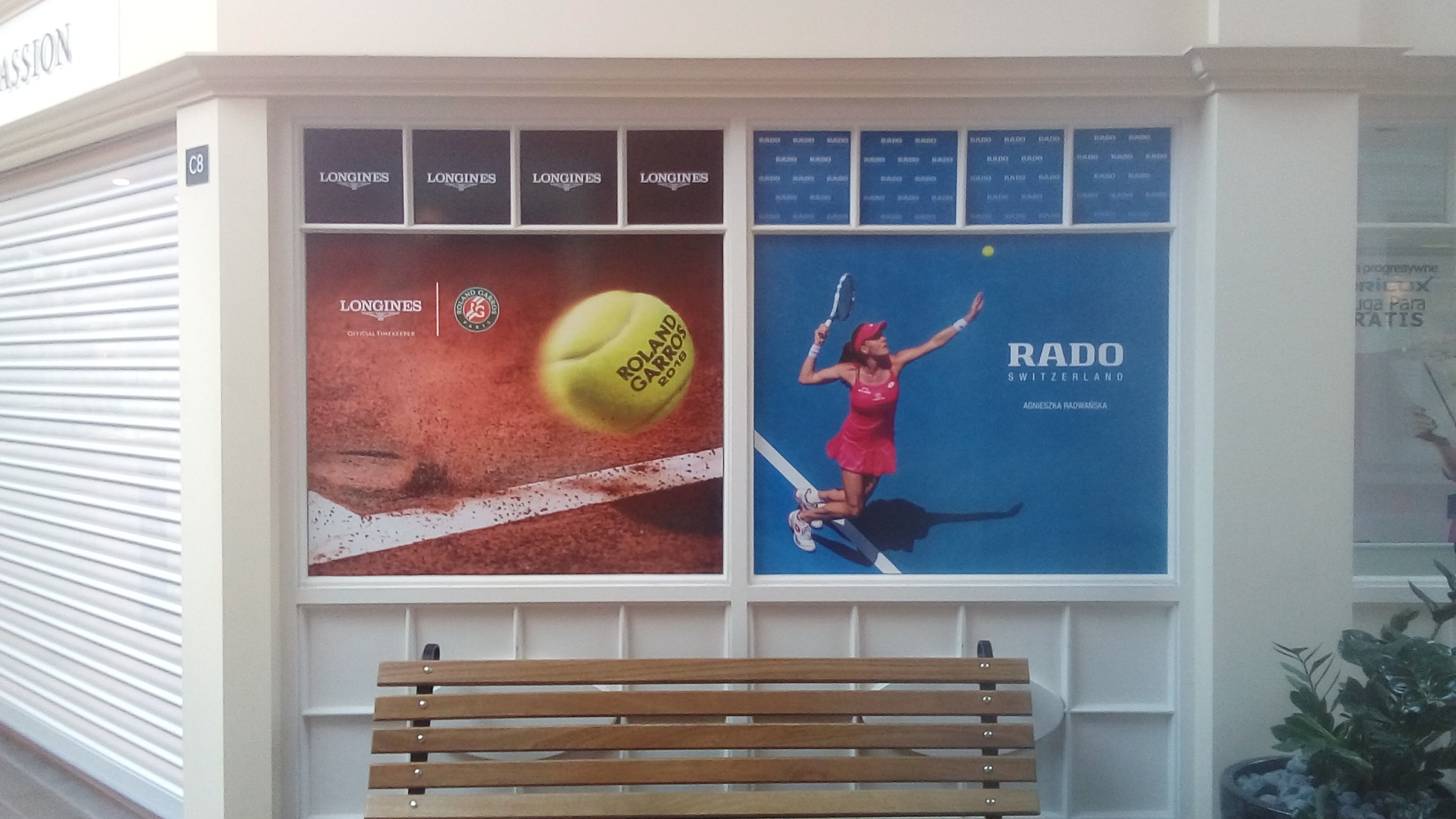 Okna witrynowe z grafikami reklamowymi turnieju Roland Garros i marki Rado, widoczne z poziomu drewnianej ławki w galerii handlowej.