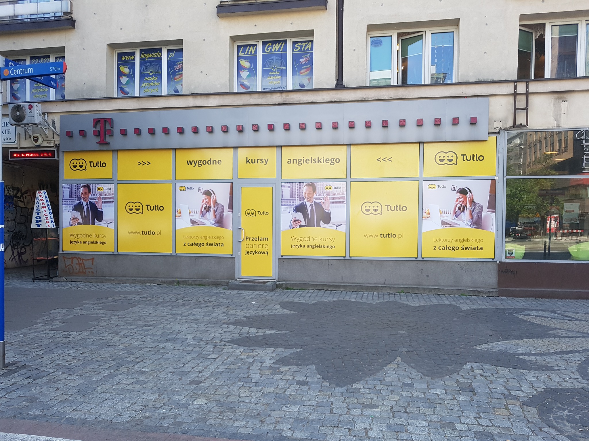 Fasada budynku z żółtymi panelami reklamowymi szkoły językowej Tutlo, widoczne logo T-Mobile na górze budynku oraz okna z reklamami innej szkoły językowej na wyższych piętrach.
