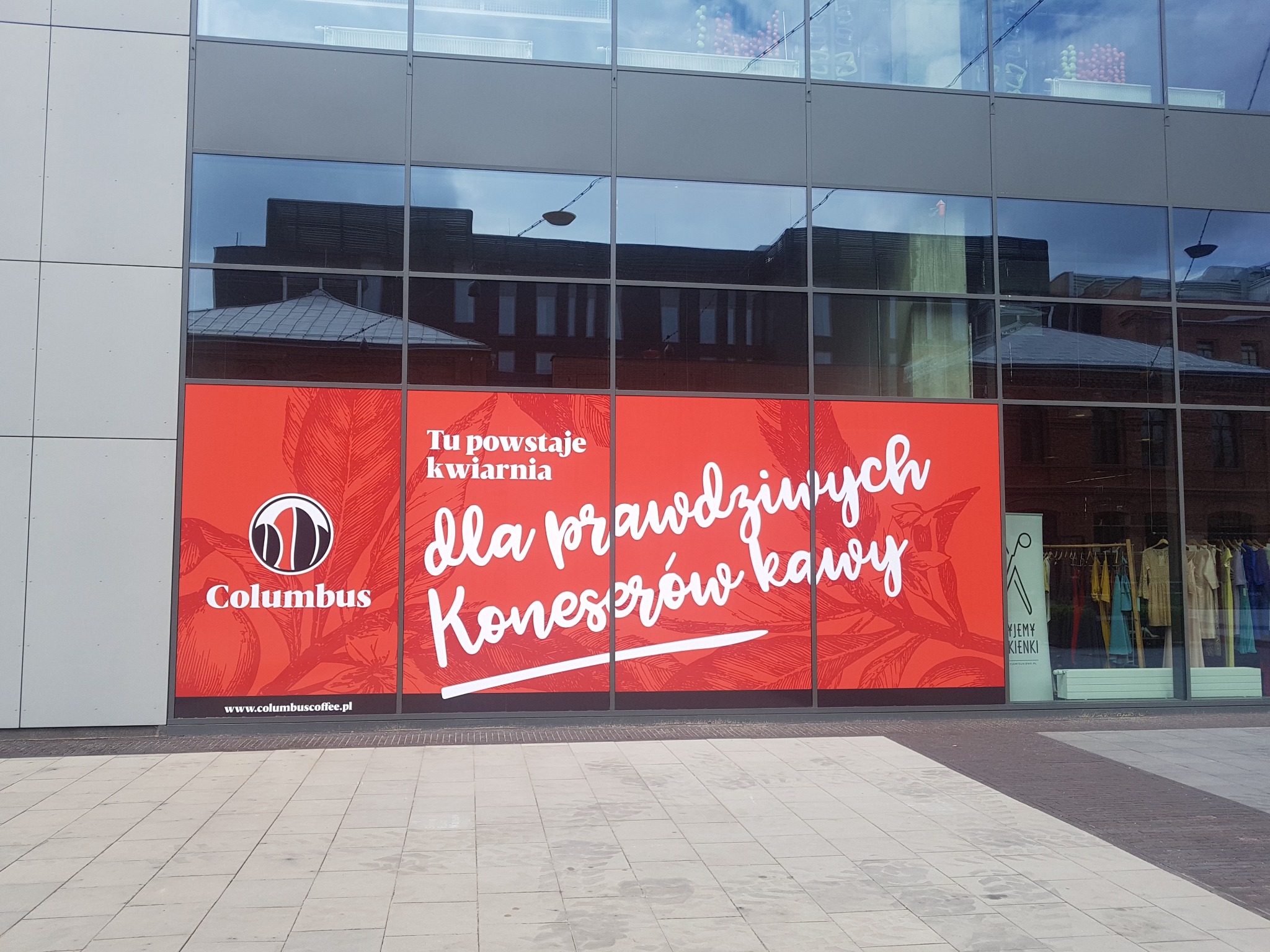 Duży, czerwony baner z napisem 'Tu powstaje kwiarnia dla prawdziwych koneserów kawy' i logo Columbus Coffee, naklejony na witrynie lokalu