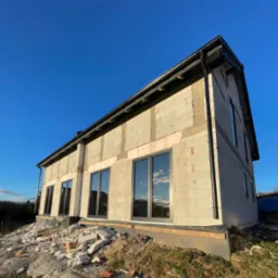 Surowy stan budynku z szarej cegły z zamontowanymi ciemnymi oknami, widoczne pozostałości materiałów budowlanych na ziemi, błękitne niebo