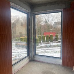 PROCARTS SPÓŁKA Z OGRANICZONĄ ODPOWIEDZIALNOŚCIĄ - Surowy stan budynku z dużymi oknami z czarnymi ramami, widok na zaśnieżony ogród z czerwoną altaną.