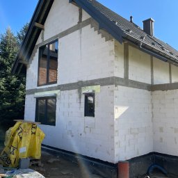 PROCARTS SPÓŁKA Z OGRANICZONĄ ODPOWIEDZIALNOŚCIĄ - Budowa domu jednorodzinnego w trakcie: wstawione okna z czarnymi ramami, ściany z białych bloczków, czarny dach z kominami, widoczne materiały budowlane i narzędzia.