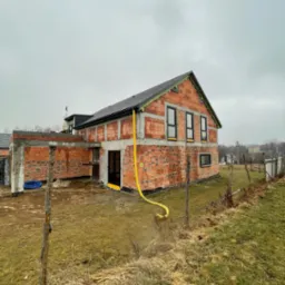 Budowa domu jednorodzinnego z oknami w czarnych ramach, widoczna instalacja żółtego węża doprowadzającego wodę.