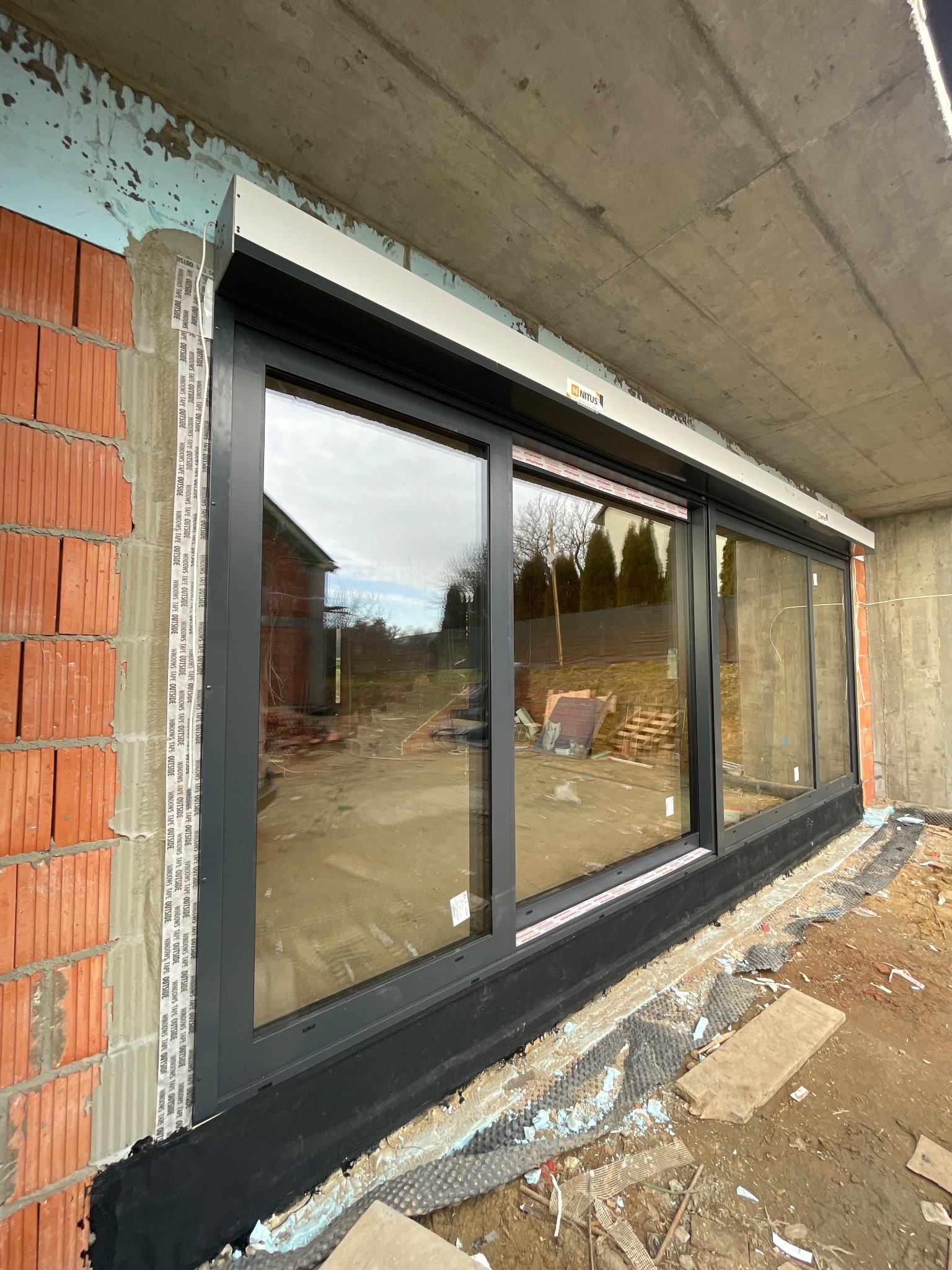 Montaż dużych, przesuwnych okien z roletami zewnętrznymi w surowym stanie budynku, widoczne cegły i taśmy uszczelniające.