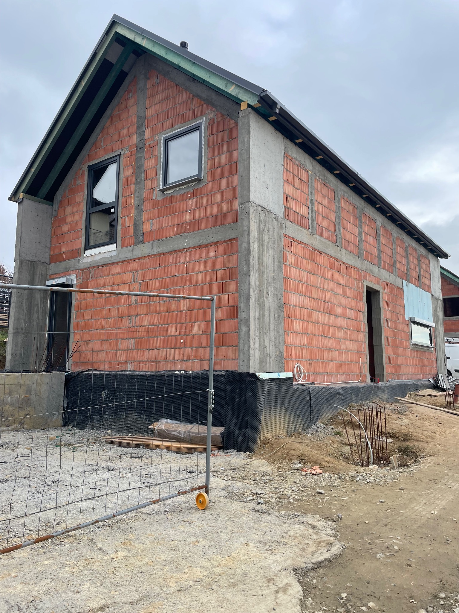 Budynek w trakcie budowy z widocznymi oknami z czarnymi ramami, ściany z czerwonej cegły, szarym betonem i czarną folią hydroizolacyjną, zabezpieczony metalowym ogrodzeniem.