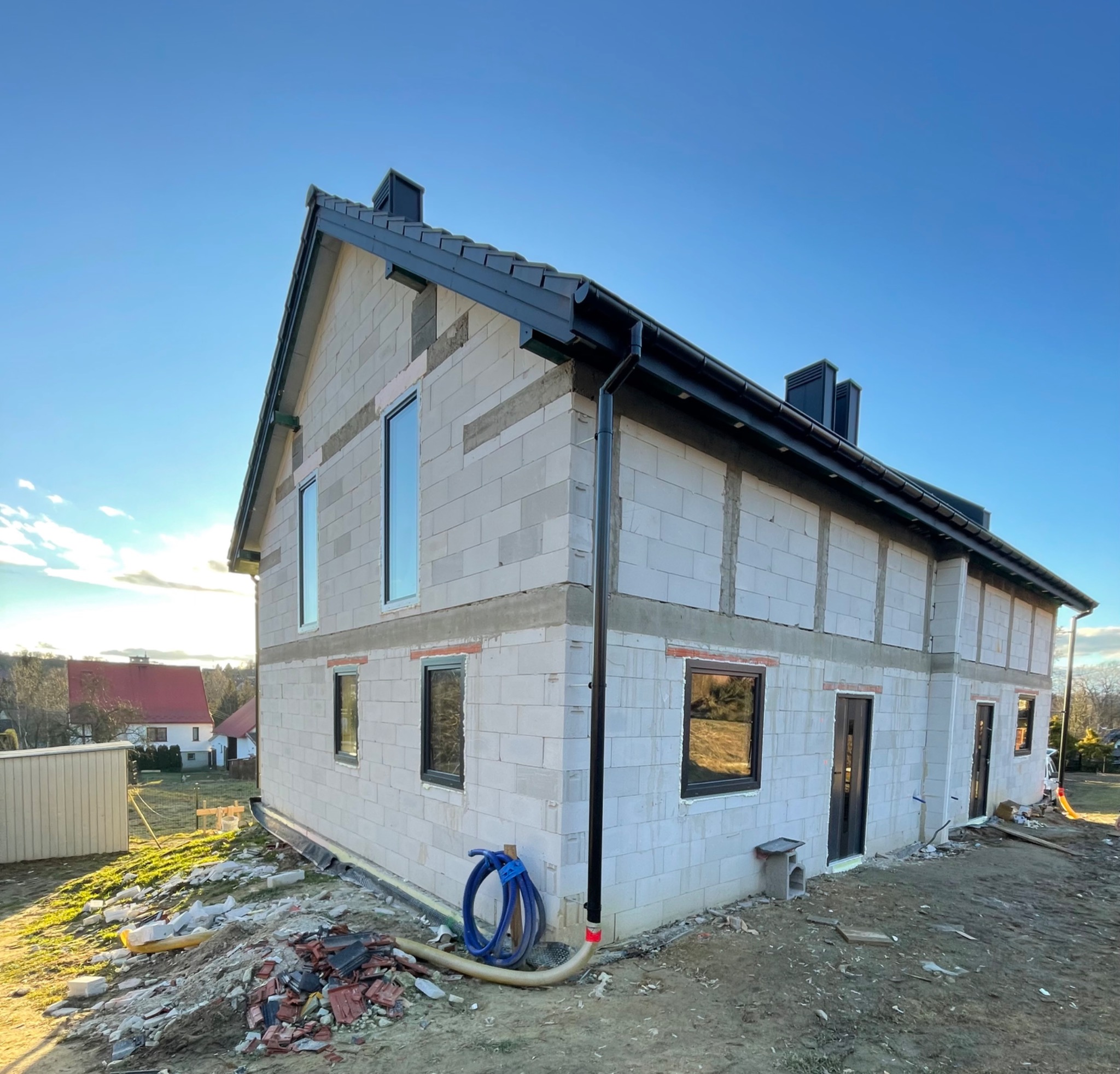 Surowy budynek z pustaków z zamontowanymi czarnymi oknami i drzwiami, widok z zewnątrz na placu budowy z niebieskim wężem leżącym obok ściany.