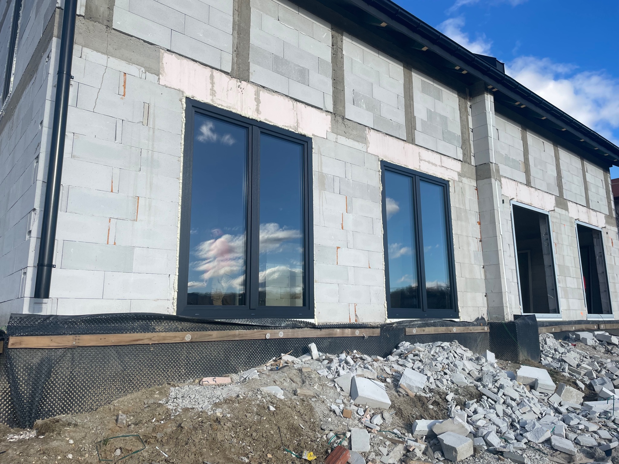 Montaż czarnych okien PCV w surowym budynku z białego betonu komórkowego, widoczne gruz i folia fundamentowa.