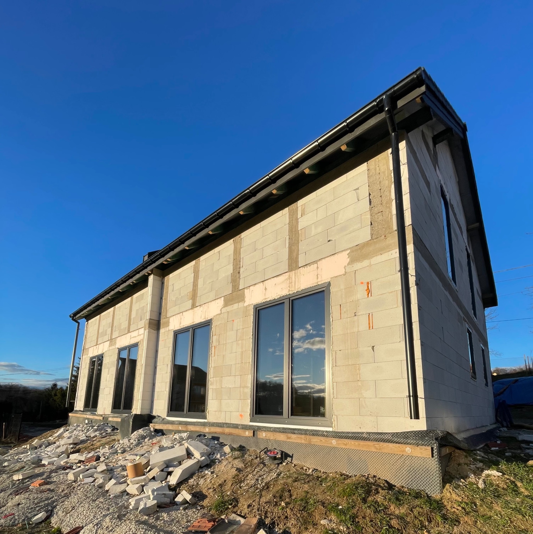 Surowy stan budynku z szarej cegły z zamontowanymi ciemnymi oknami, widoczne pozostałości materiałów budowlanych na ziemi, błękitne niebo