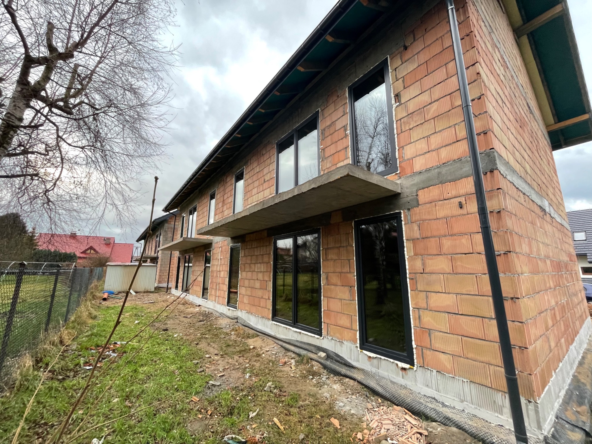 Budynek w trakcie budowy z zamontowanymi czarnymi oknami, widoczne surowe cegły i elementy konstrukcyjne, pochmurne niebo w tle.