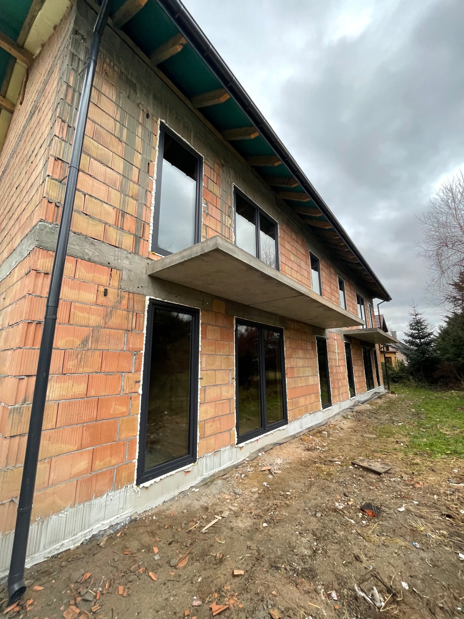 Surowy budynek w trakcie montażu stolarki okiennej, widoczne czarne ramy okienne i surowa elewacja z cegły, na zewnątrz teren budowy z ziemią i resztkami materiałów.
