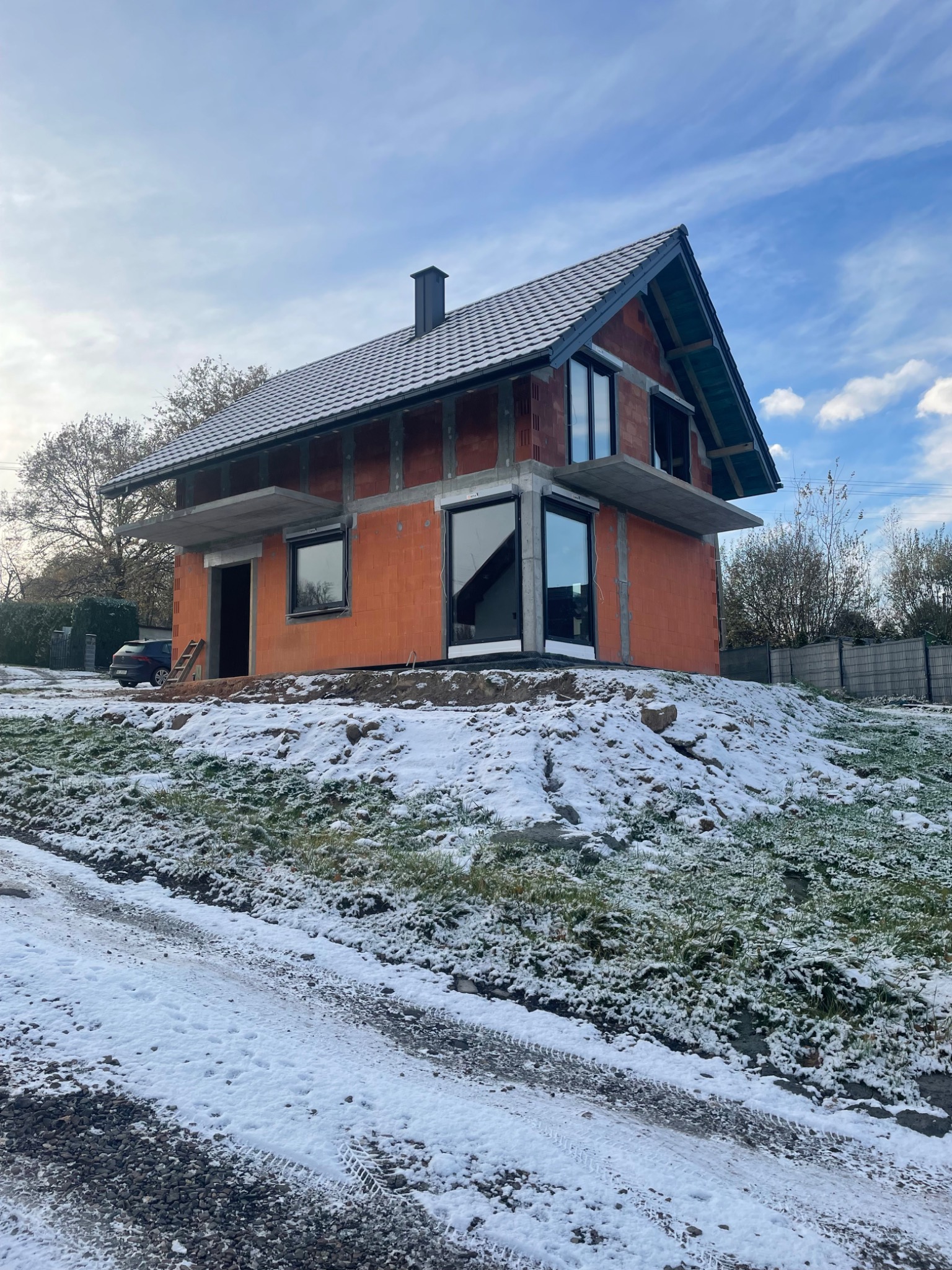 Surowy budynek w stanie deweloperskim z zamontowanymi oknami, częściowo ośnieżony, widok z dołu na tle pochmurnego nieba.