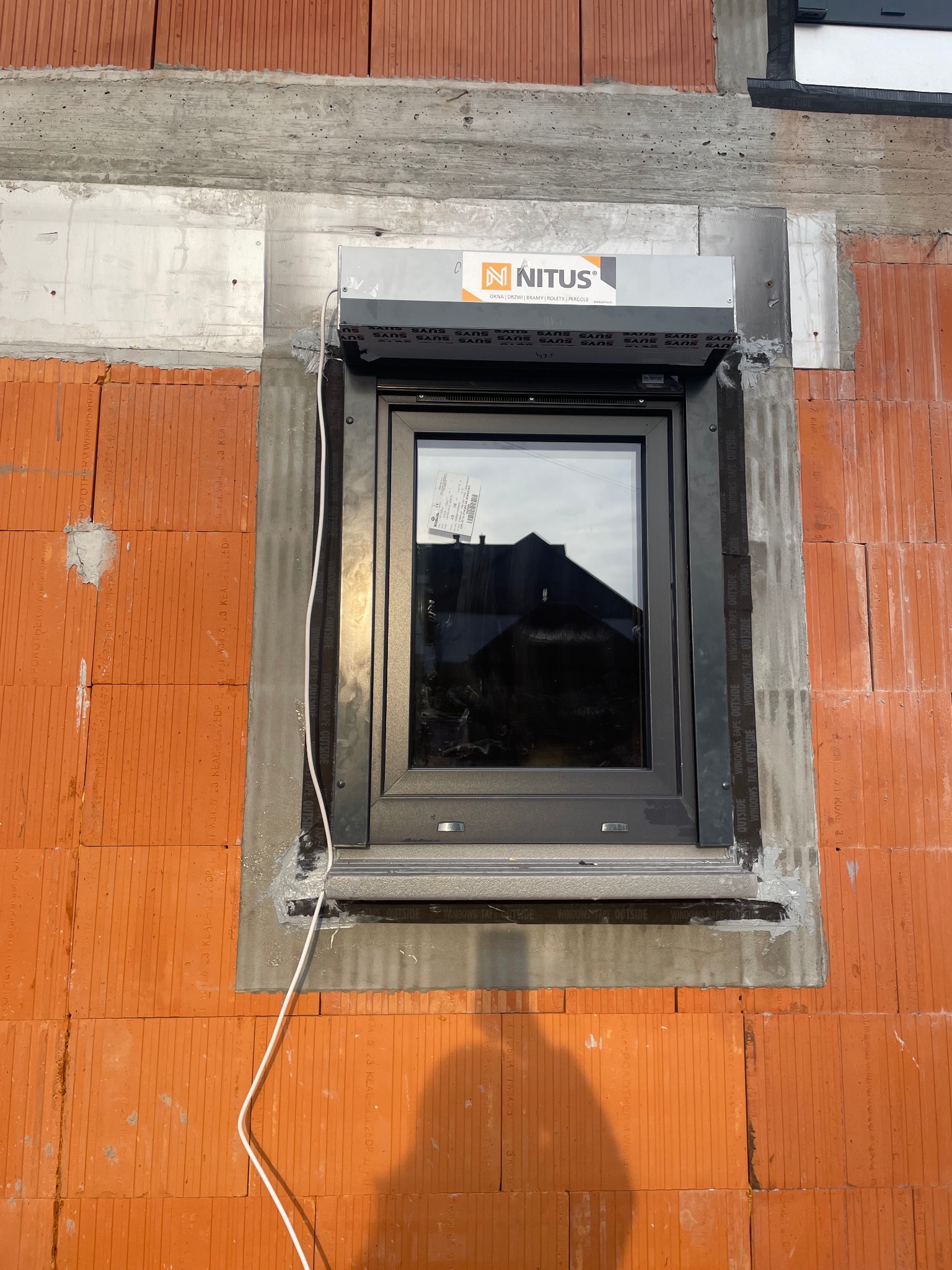 Montaż okna PCV z roletą zewnętrzną w nowym budynku z czerwonej cegły, widoczne taśmy uszczelniające i logo Nitus.