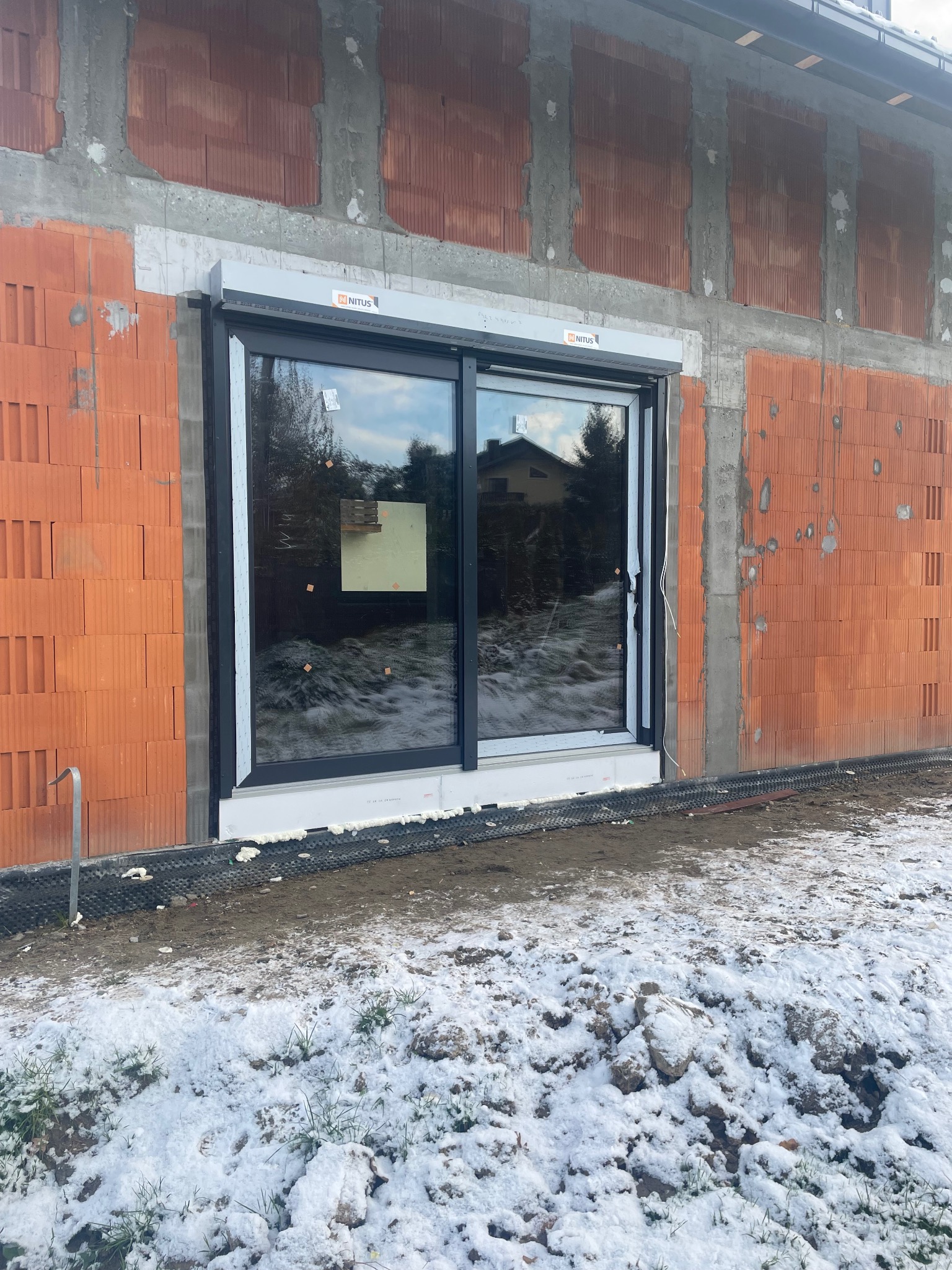 Montaż okna przesuwnego z roletą zewnętrzną w surowym stanie budynku, widoczne bloczki ceramiczne i śnieg na ziemi.
